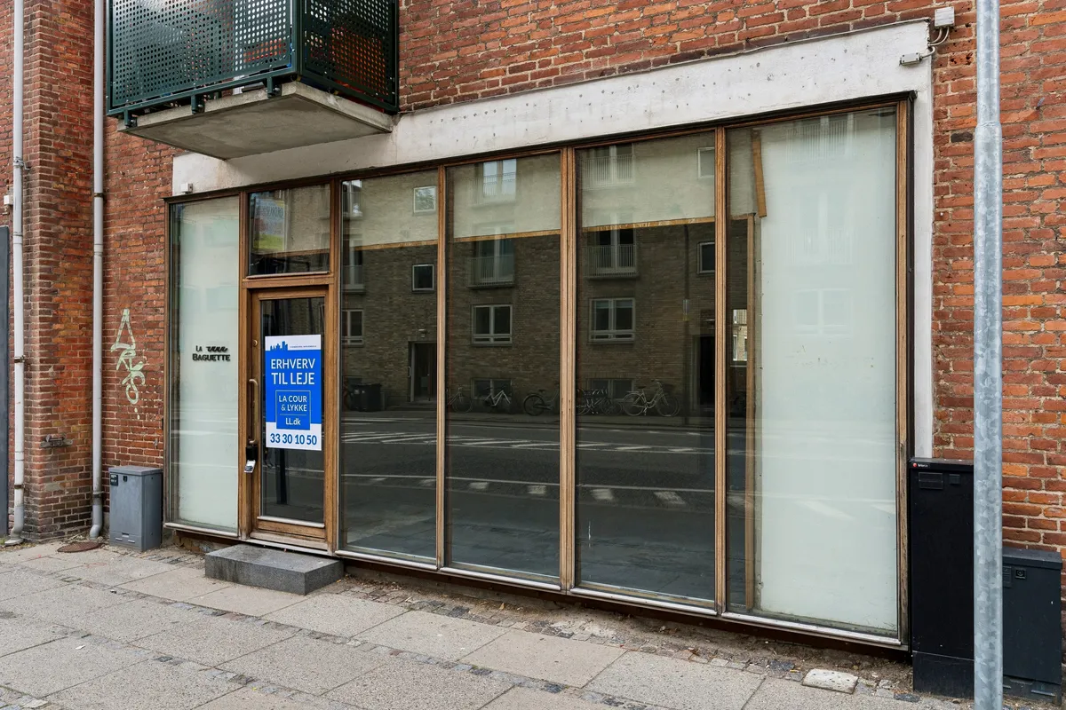 Lejlighed til leje på Ordrupvej 62B i Charlottenlund med slanke glasvinduer og skilte med tekst på dansk. Bygningen er rødmalet og har en balkon og skraldespand.