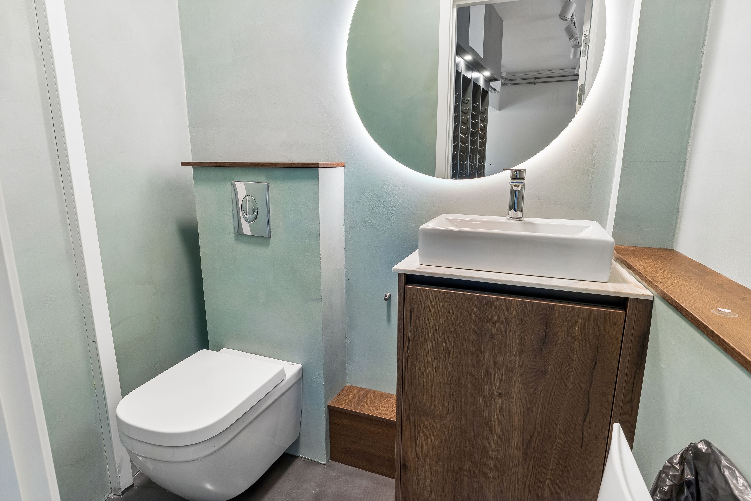 Rundt spejl og høj gulvskabsdør i badeværelset med hvidt toilet og vask samt håndklædestang med håndklæde