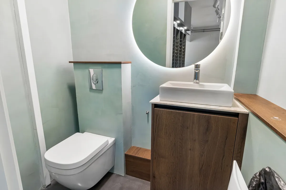 Rundt spejl og høj gulvskabsdør i badeværelset med hvidt toilet og vask samt håndklædestang med håndklæde