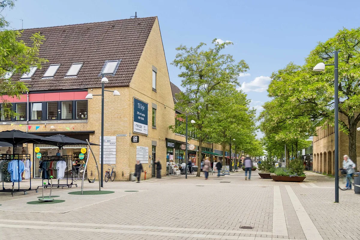 Gågade med solbeskyttelse, tøjforretning, cykler og gående folk i Allerød Bymidte med smukke træer, planter og solrige veje