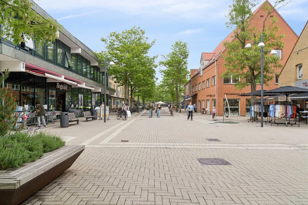På gågaden i Allerød Bymidte er der en bygning med butikker og en bred gågade med folk, der går og cykler langs bredden