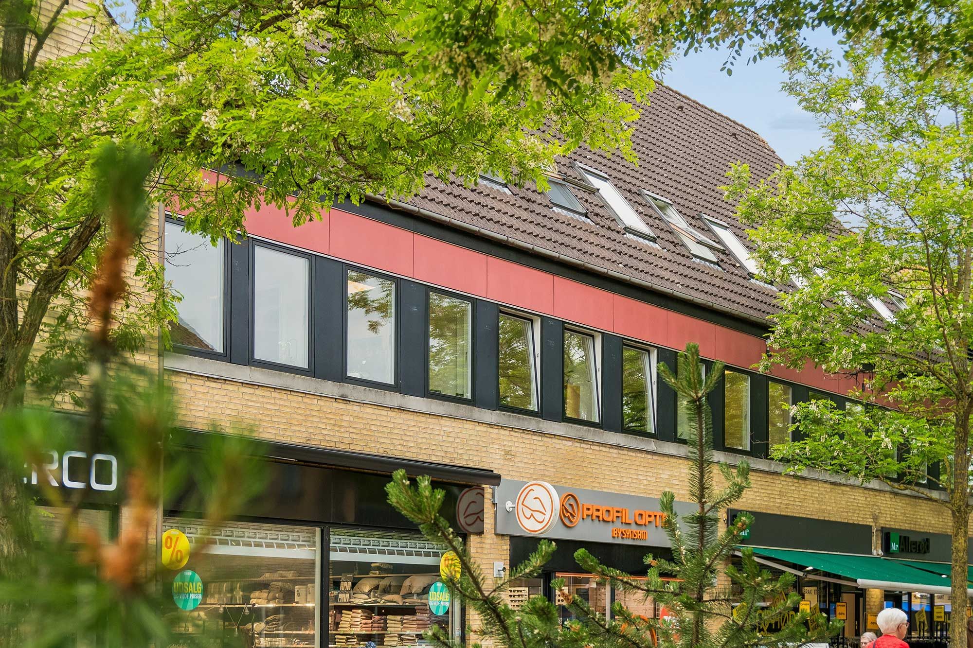 Kontor- eller klinikbygning i Allerød Bymidte med mange vinduer, røde gavle og et stort skrift Profil Optik Bysviidsen. En mand og en kvinde går forbi bygningen