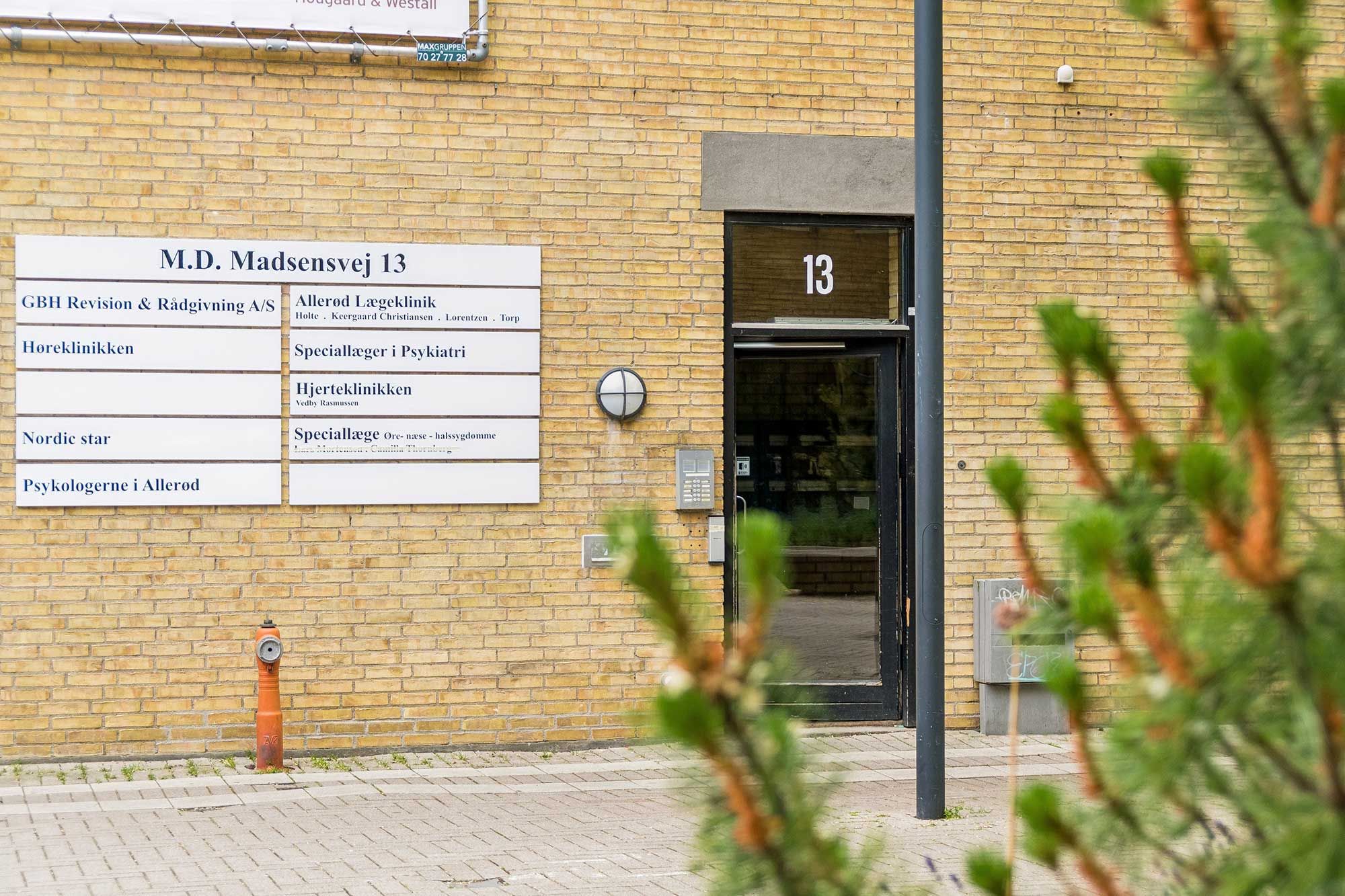 Bygning med glasdøre og skilte med M.D. Madsensvej 13, Høreklinikken, Psykologerne i Allerød og Speciallæger i Psykiatri. Brændenælde og brandvandsslange ved siden af.