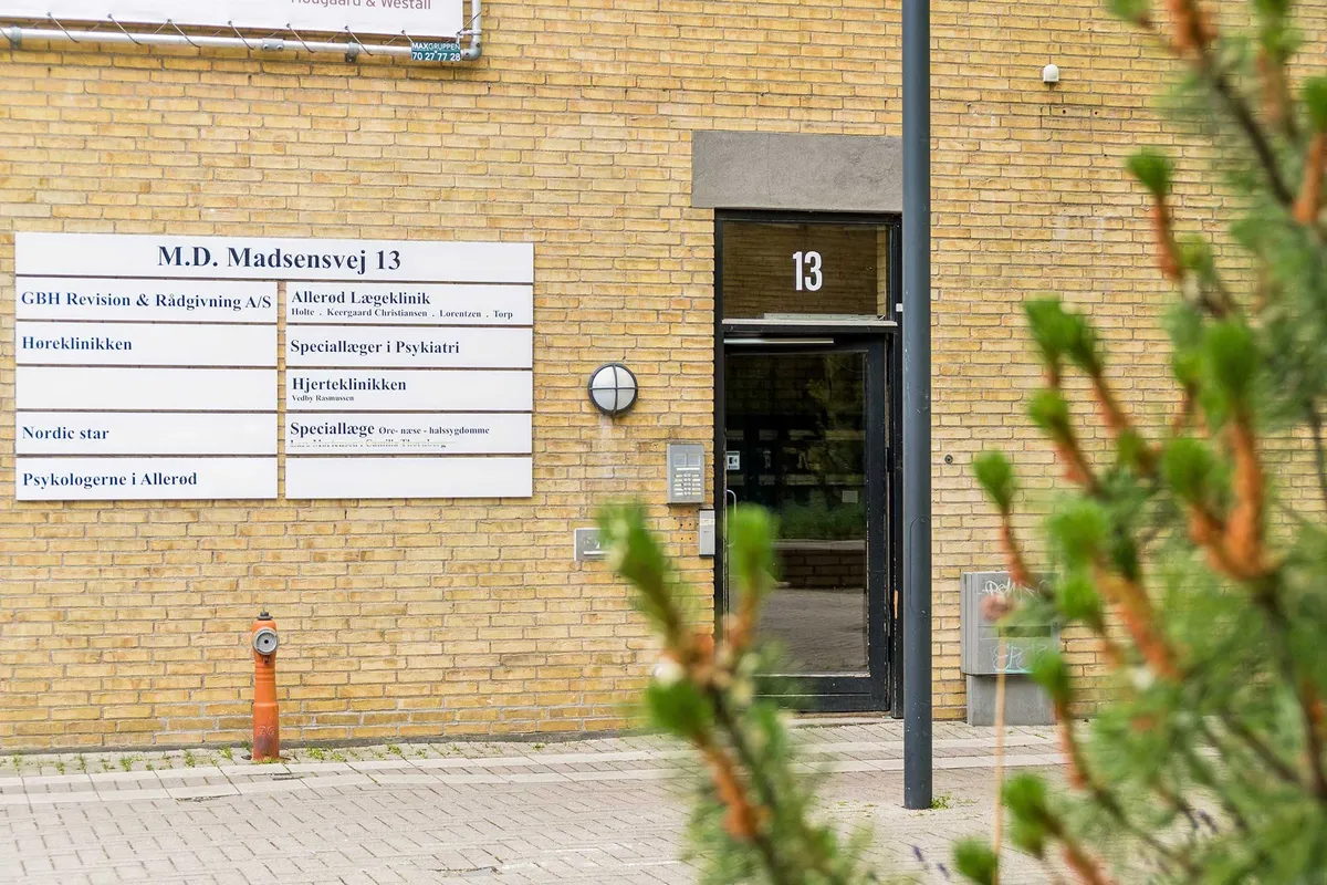 Bygning med glasdøre og skilte med M.D. Madsensvej 13, Høreklinikken, Psykologerne i Allerød og Speciallæger i Psykiatri. Brændenælde og brandvandsslange ved siden af.