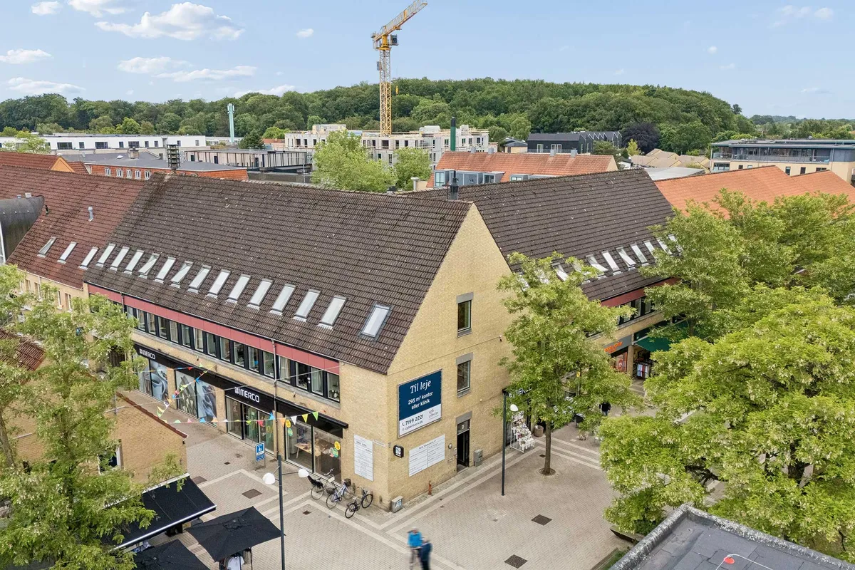 En klinik i Allerød Bymidte med røde tag og parkeringsmuligheder. Bygningen har flere vinduer, og der er grønne træer foran. I baggrunden er der flere huse og en kran.
