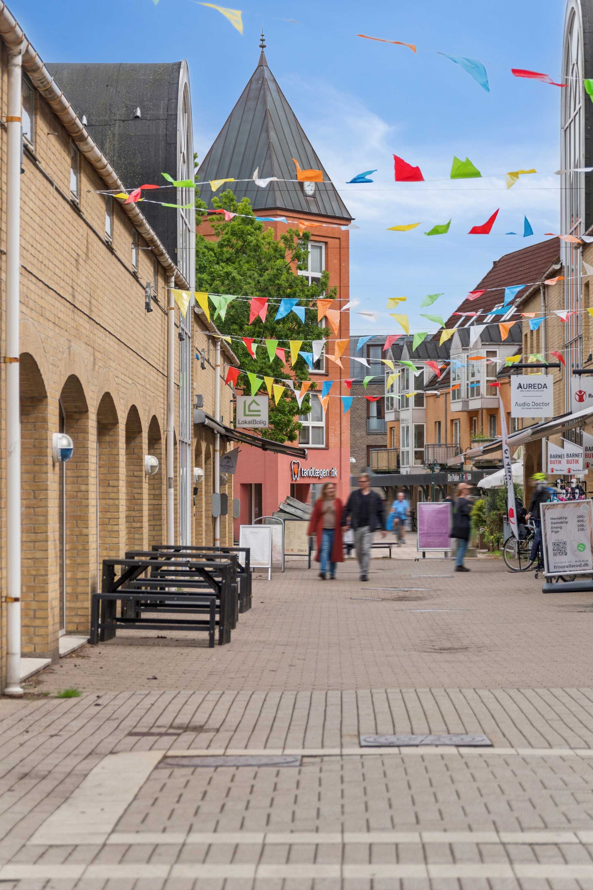En gågade i Allerød Bymidte med smukke farverige flag og mennesker i bevægelse, slanke bygninger og slanke bænke