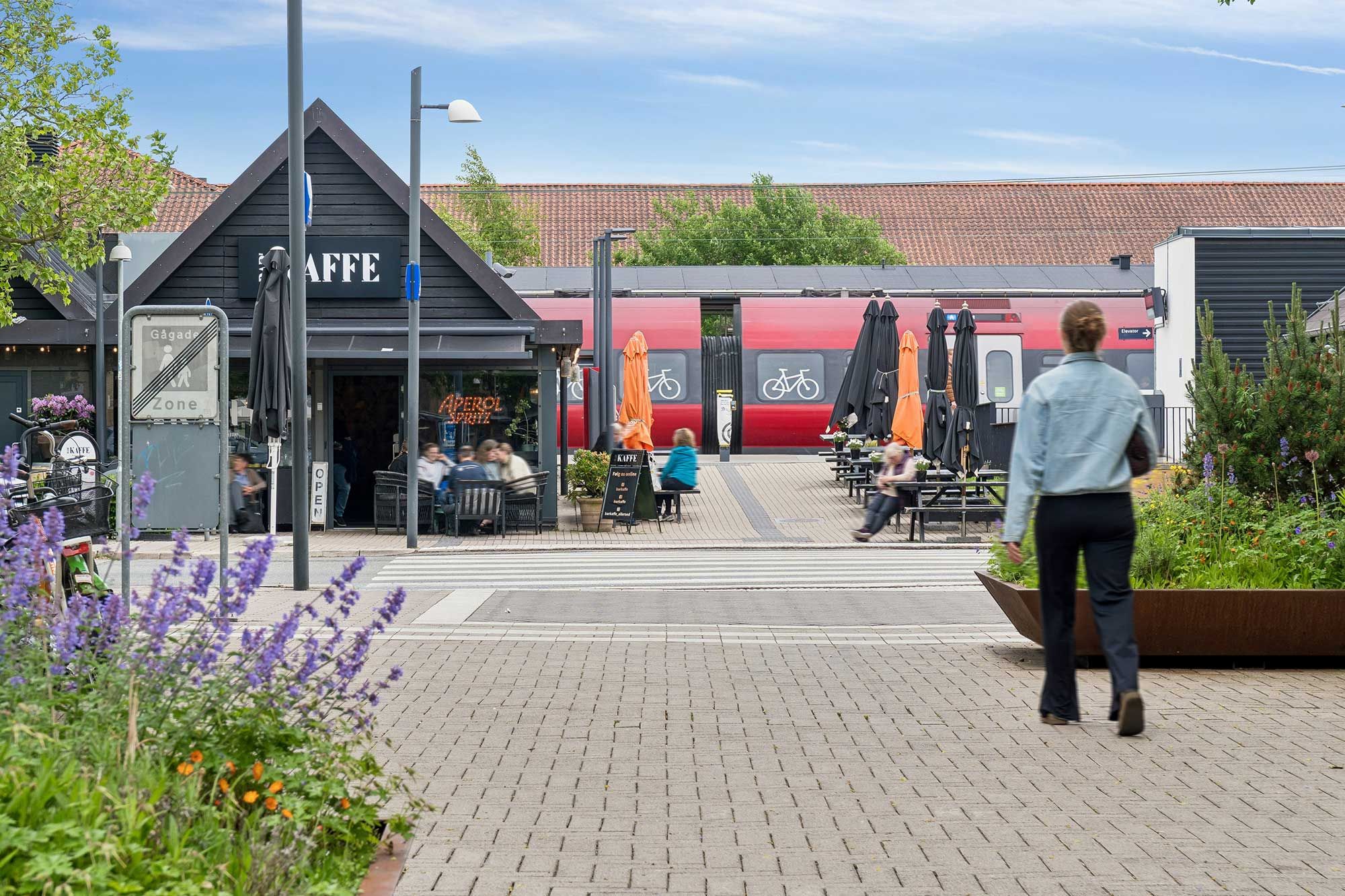En voksen kvinde går mod en togstation med caféer, blomsterbed og en vejledningstavle i Allerød Bymidte