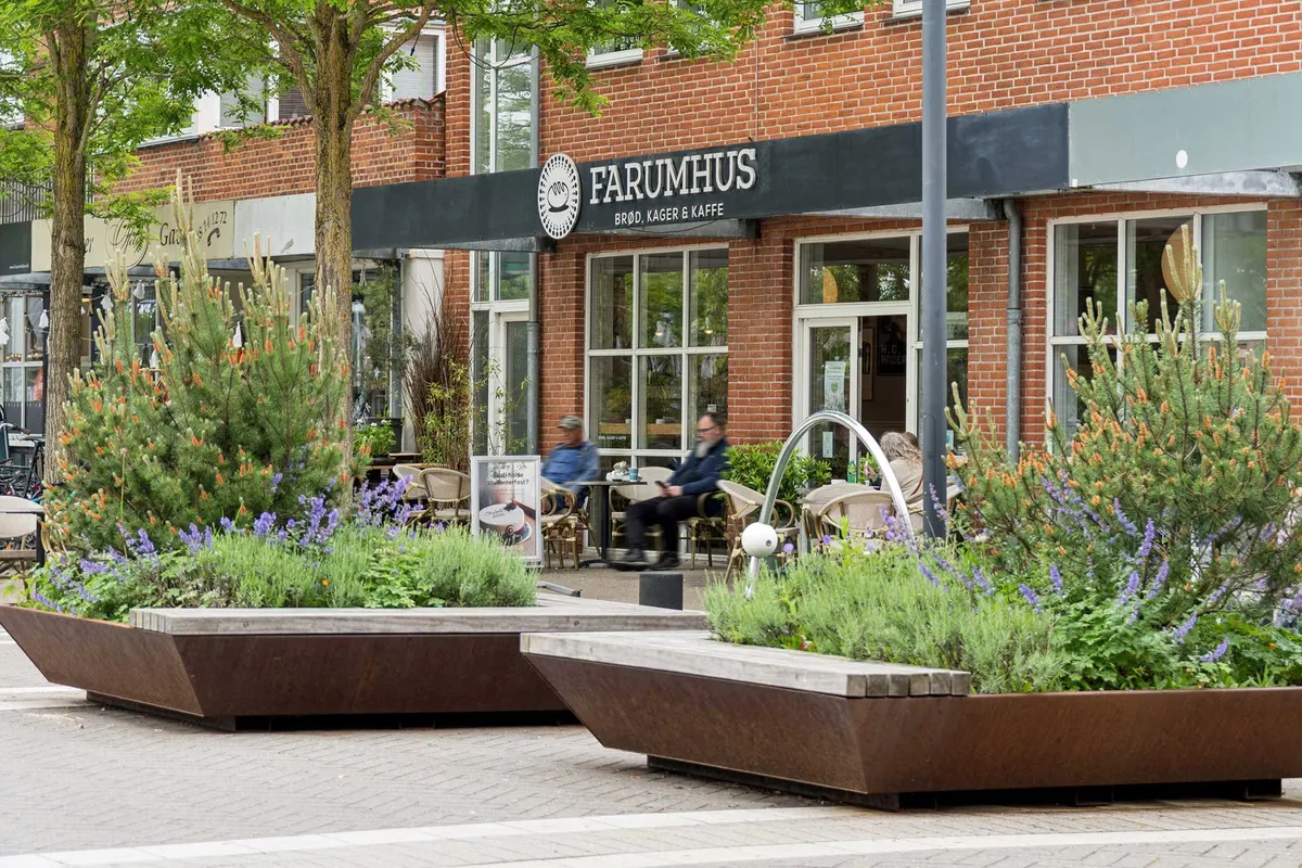 Farumhus café med lyserøde planter og lyserøde blomster i forgrunden og to stolpebord med gæster og søjler i baggrunden
