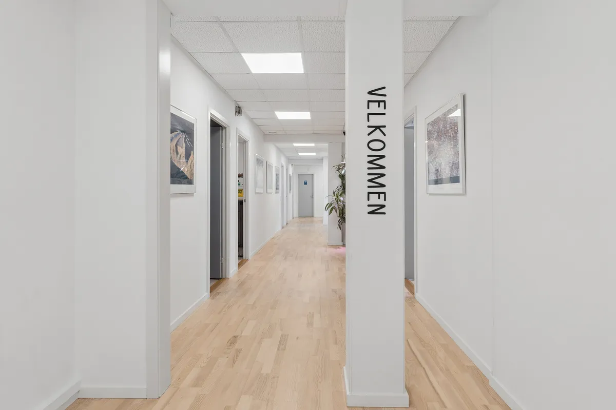 Indersiden af et hvidt kontorlokale med lyse lofter og lysepunkter. Hallen er udstyret med lysegrå vægge og lysegule gulve. Der er en søjle med teksten Velkommen, og der er flere døre med vægmalerier