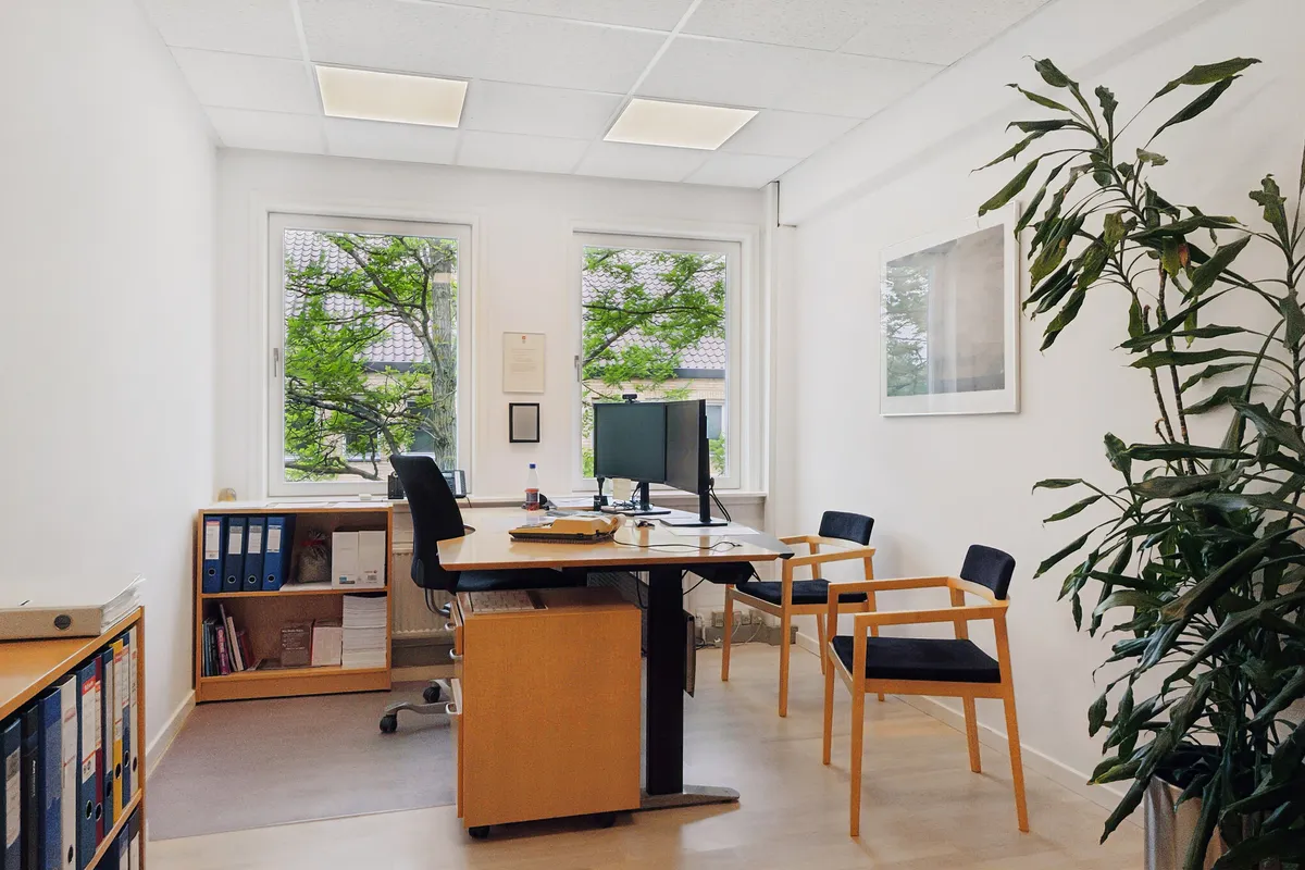 Kontorrum med moderne møbler og teknologi samt trægulve, vinduer og plante.