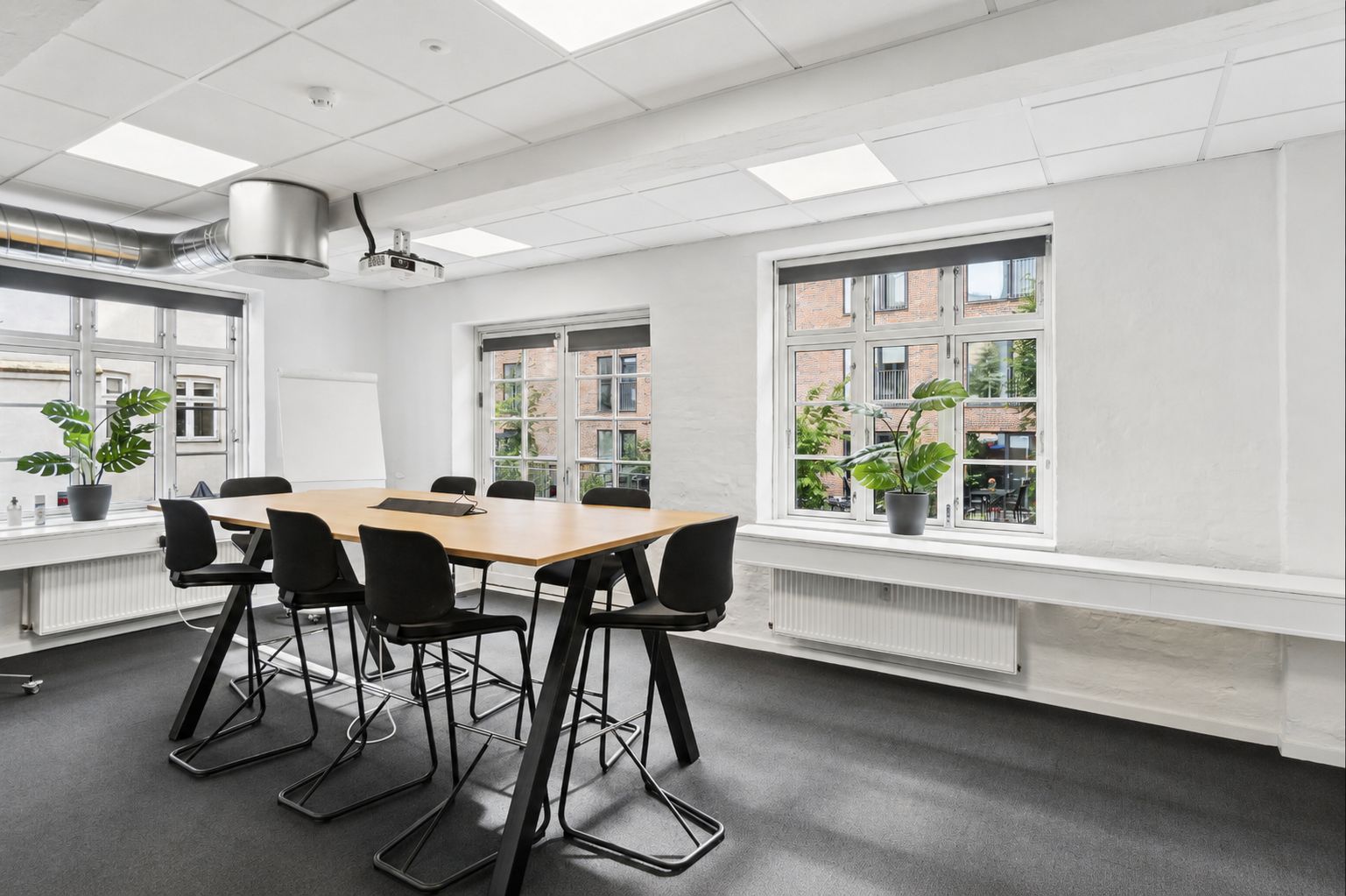Kontor med lyse loftsbjælke og lysegulv, bord og stole, plante og skrivebord, vindue med plante og radiator, projektor og ventilationsrør