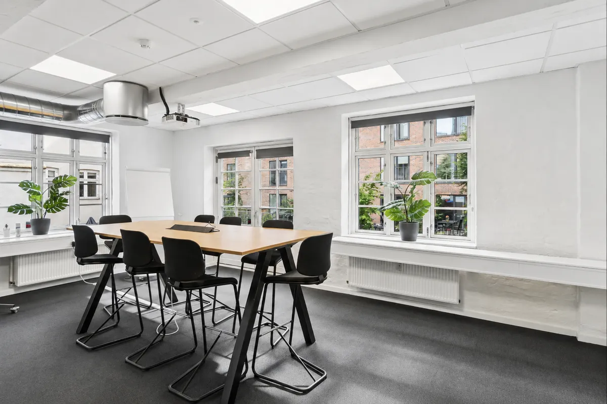 Kontor med lyse loftsbjælke og lysegulv, bord og stole, plante og skrivebord, vindue med plante og radiator, projektor og ventilationsrør