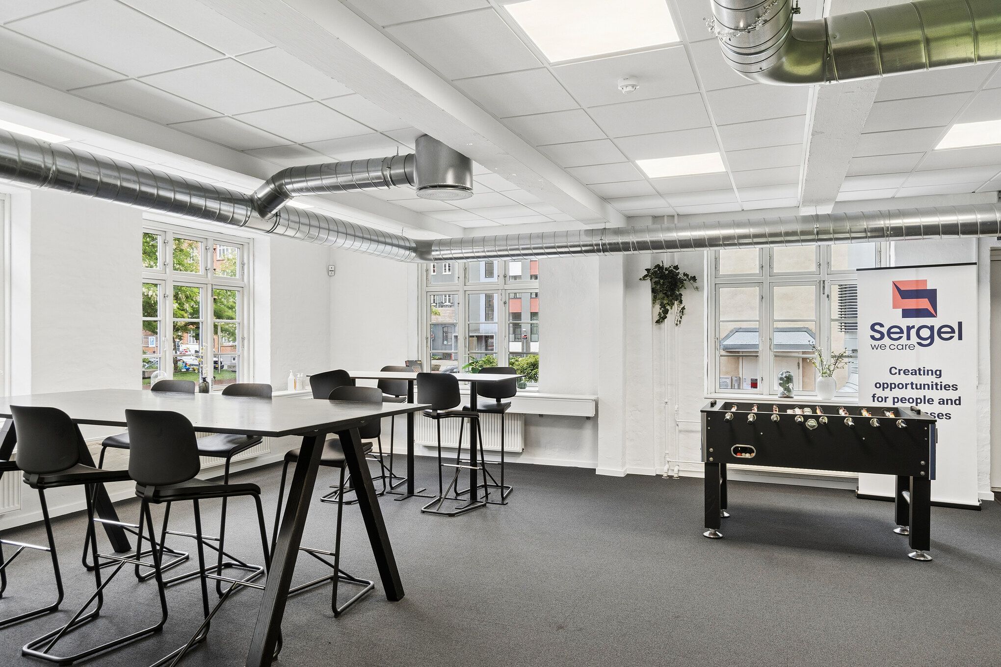 Kontor med store vinduer, højt loft og tekniske faciliteter, fælles kantine og tagterrasse, stolpebord og fodboldbord, smykket med en plante i kassen