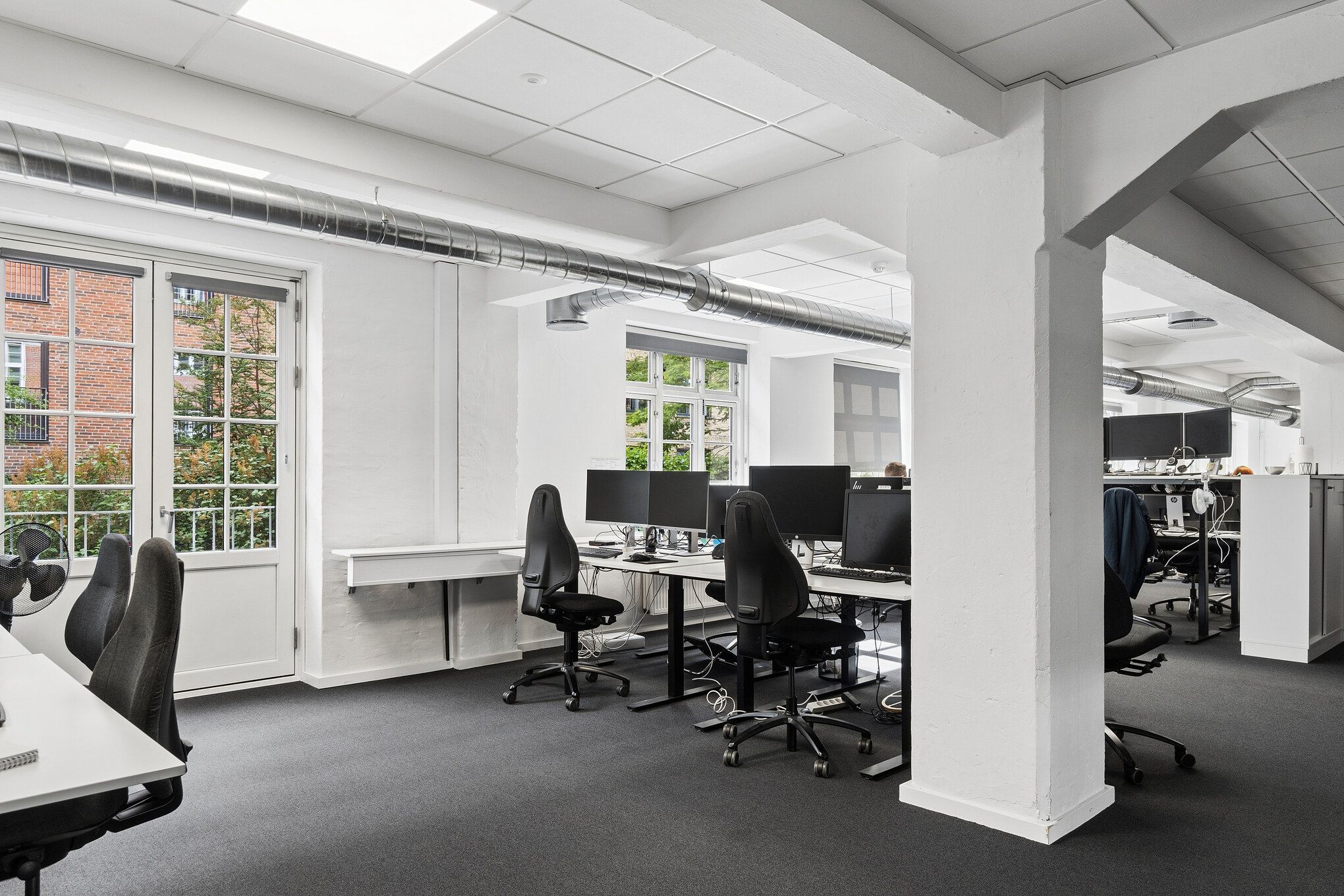 Kontorrum med høje lofter, lyse gulve og vinduer med udsigt til grønne træer. Kontorbord med monitorer, skrivebordslampe og ventilator. Høj kvalitet.