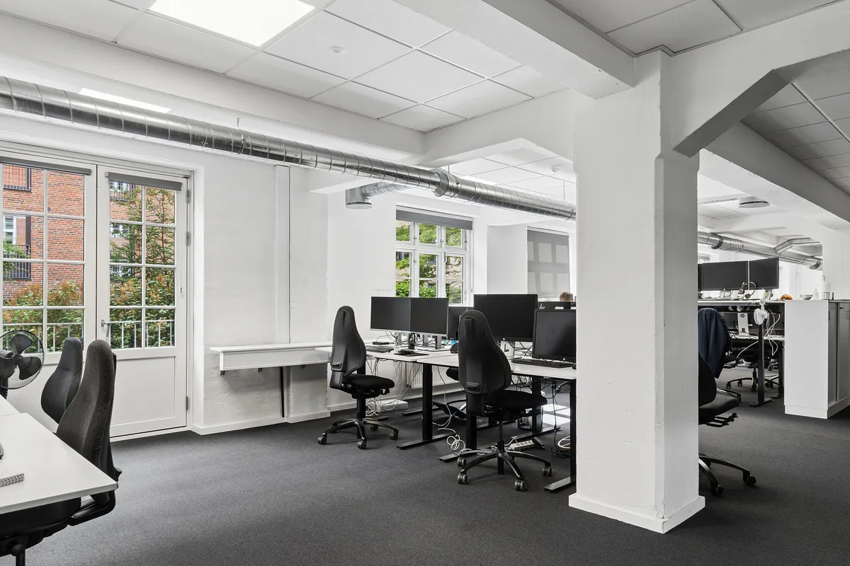 Kontorrum med høje lofter, lyse gulve og vinduer med udsigt til grønne træer. Kontorbord med monitorer, skrivebordslampe og ventilator. Høj kvalitet.