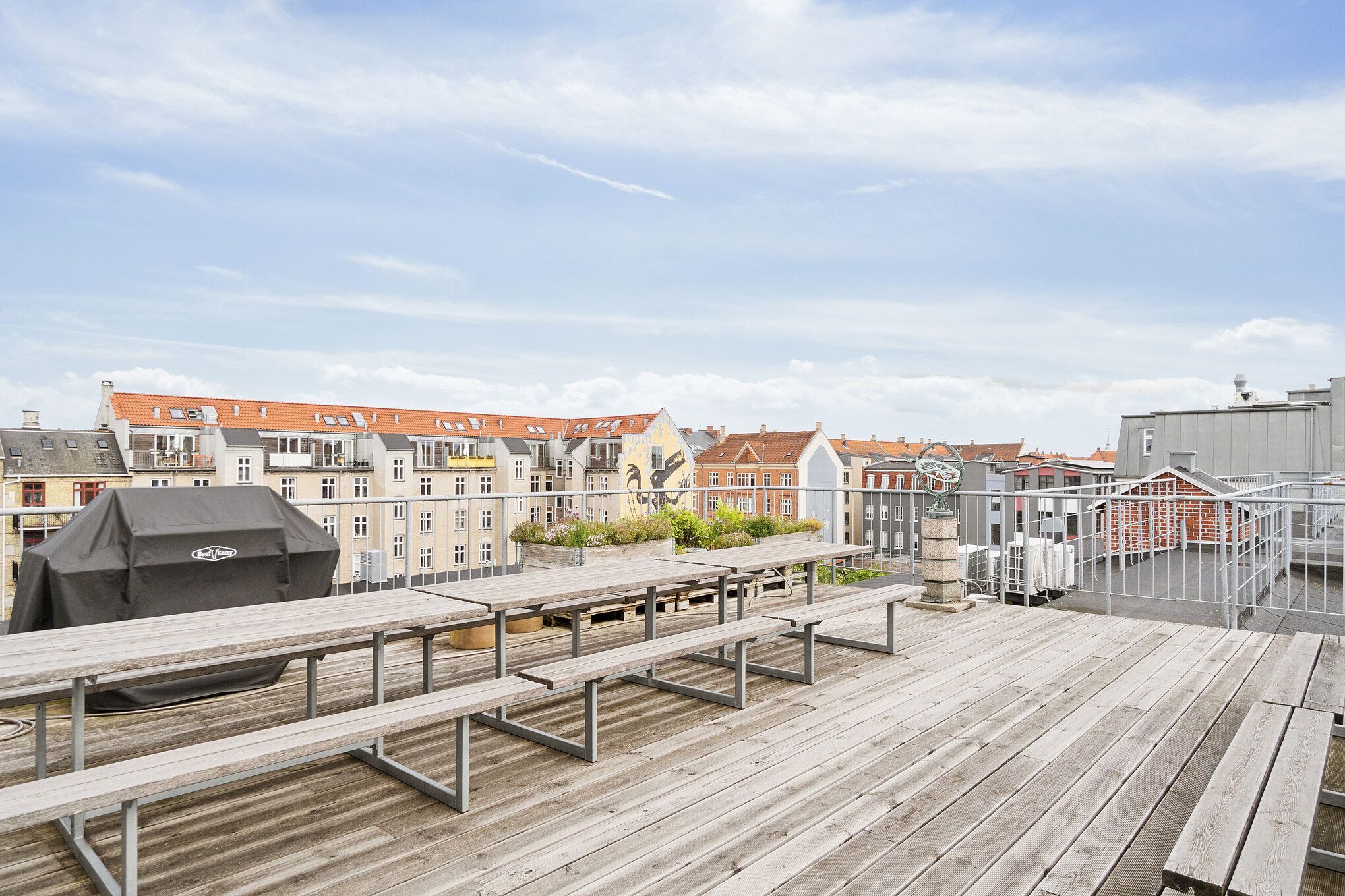 Terrasse med bord, bænke og grill. En udsigt over bygninger og en klar himmel med skyer. En skulptur i sten og et metalhegn