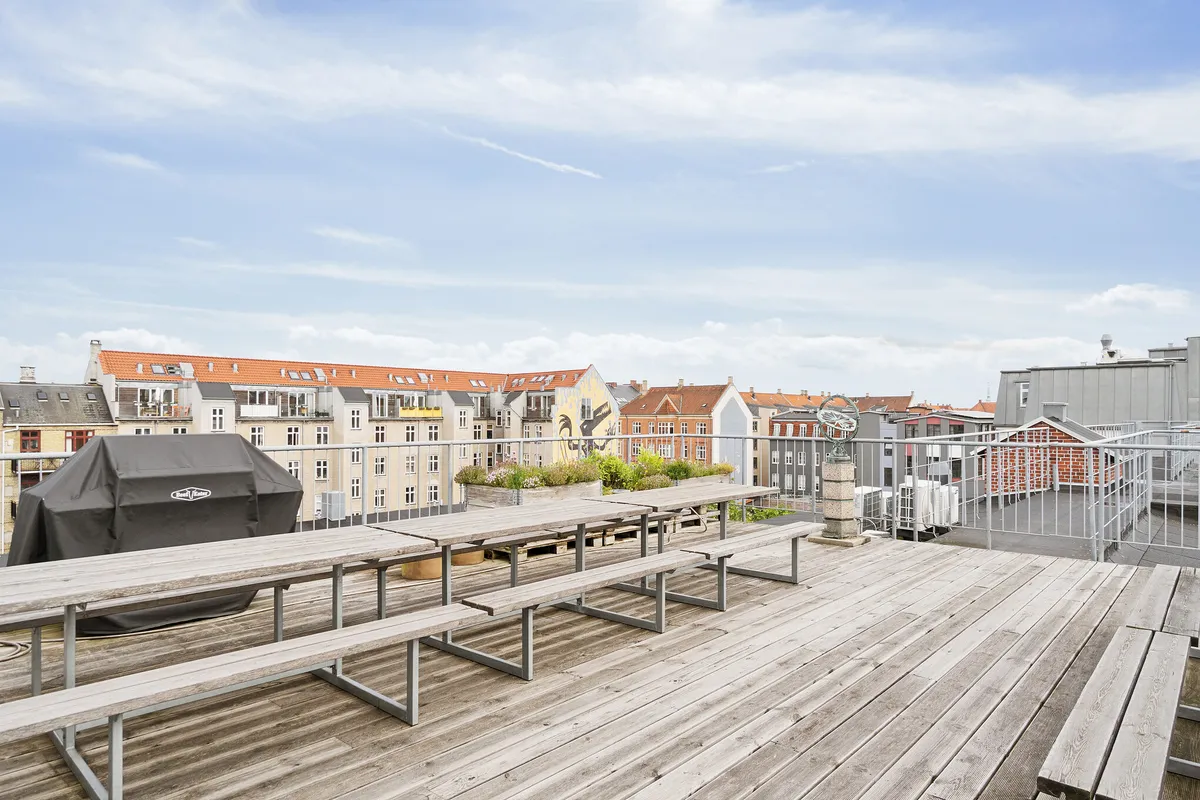 Terrasse med bord, bænke og grill. En udsigt over bygninger og en klar himmel med skyer. En skulptur i sten og et metalhegn