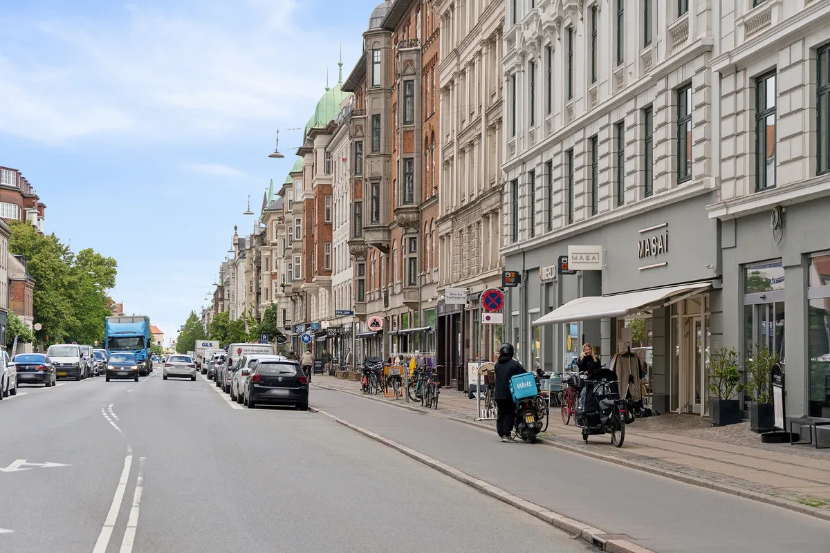 Østerbros gade med parkerede biler og forretninger, hvor flere gående og en cykel er synlige samt en vogn med hvidt logo