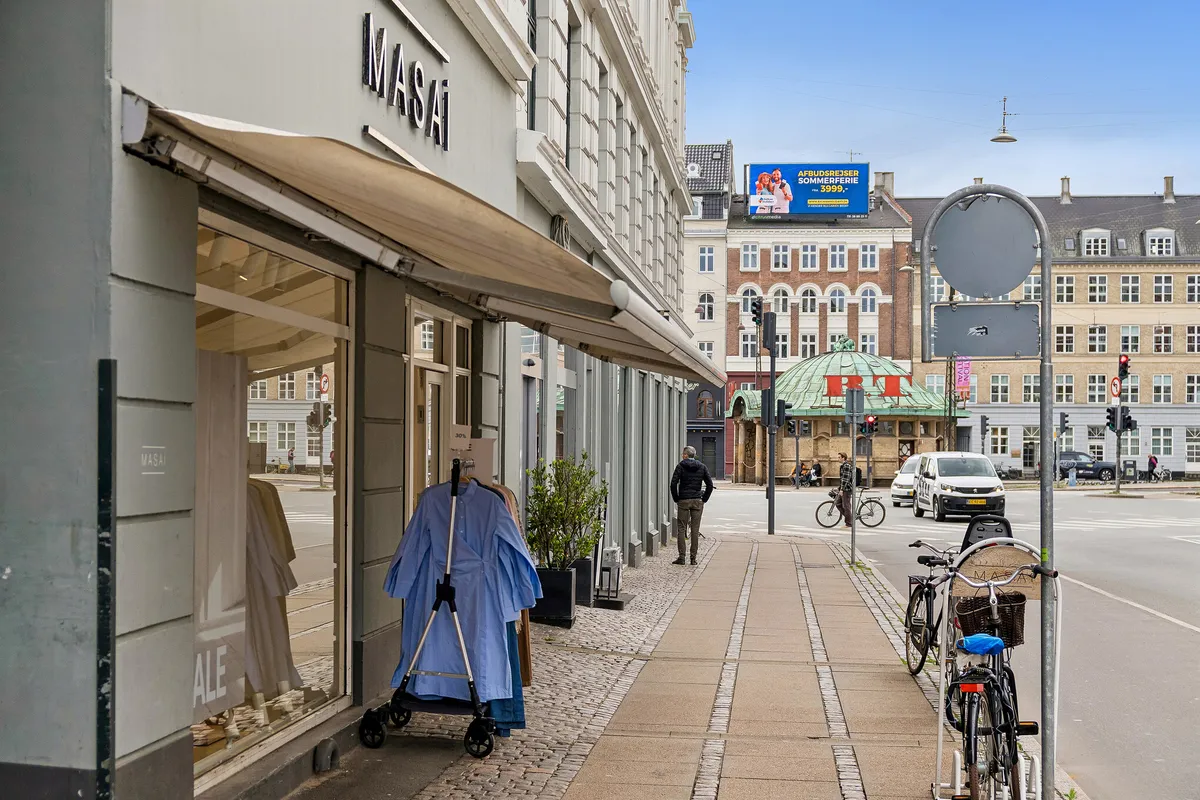 Mennesker går på fortov med cykler og kørestole. Biler og cykler er på vejen. Butikker med solskærme og reklameskilte. Børnevagtstativ og klædeskab er i butiksvinduet.