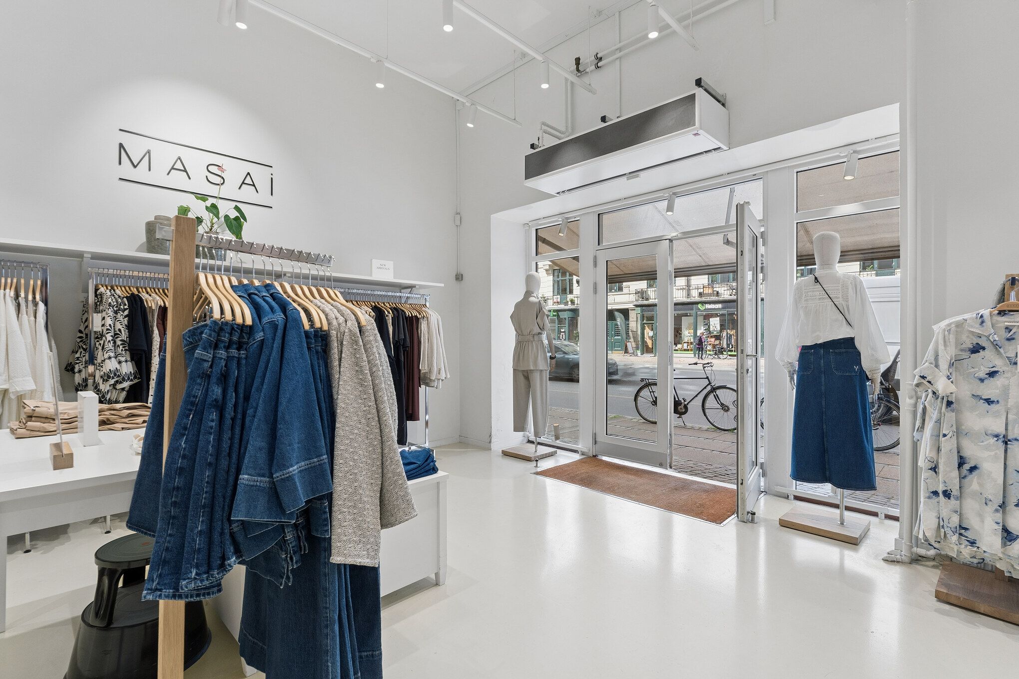 Butik med tøj og tøjstandere med mærket Masai, tøj på manequin og en plante i skudfat, samt udgang og vej med cykel og bygning med vindue i baggrunden