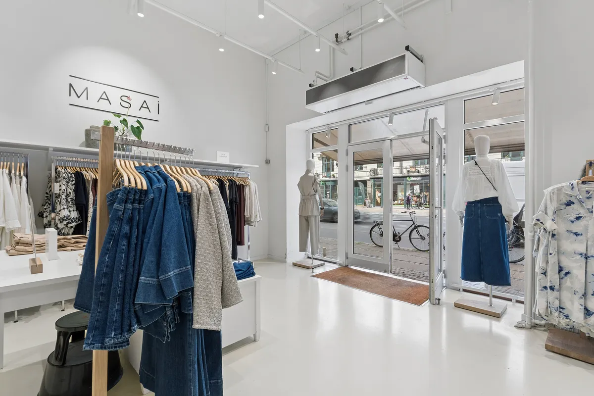 Butik med tøj og tøjstandere med mærket Masai, tøj på manequin og en plante i skudfat, samt udgang og vej med cykel og bygning med vindue i baggrunden