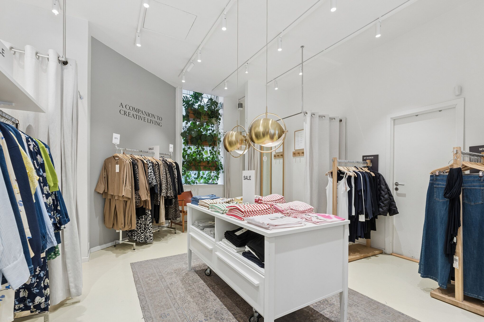 Et moderne butikslejemål med en stor, hvid midterbord, hvor tøj er udstillet og solgt. Rundt om er der hængehylder med tøj og trægulve. Der er hængende lysekroner og plantegrønne i baggrunden