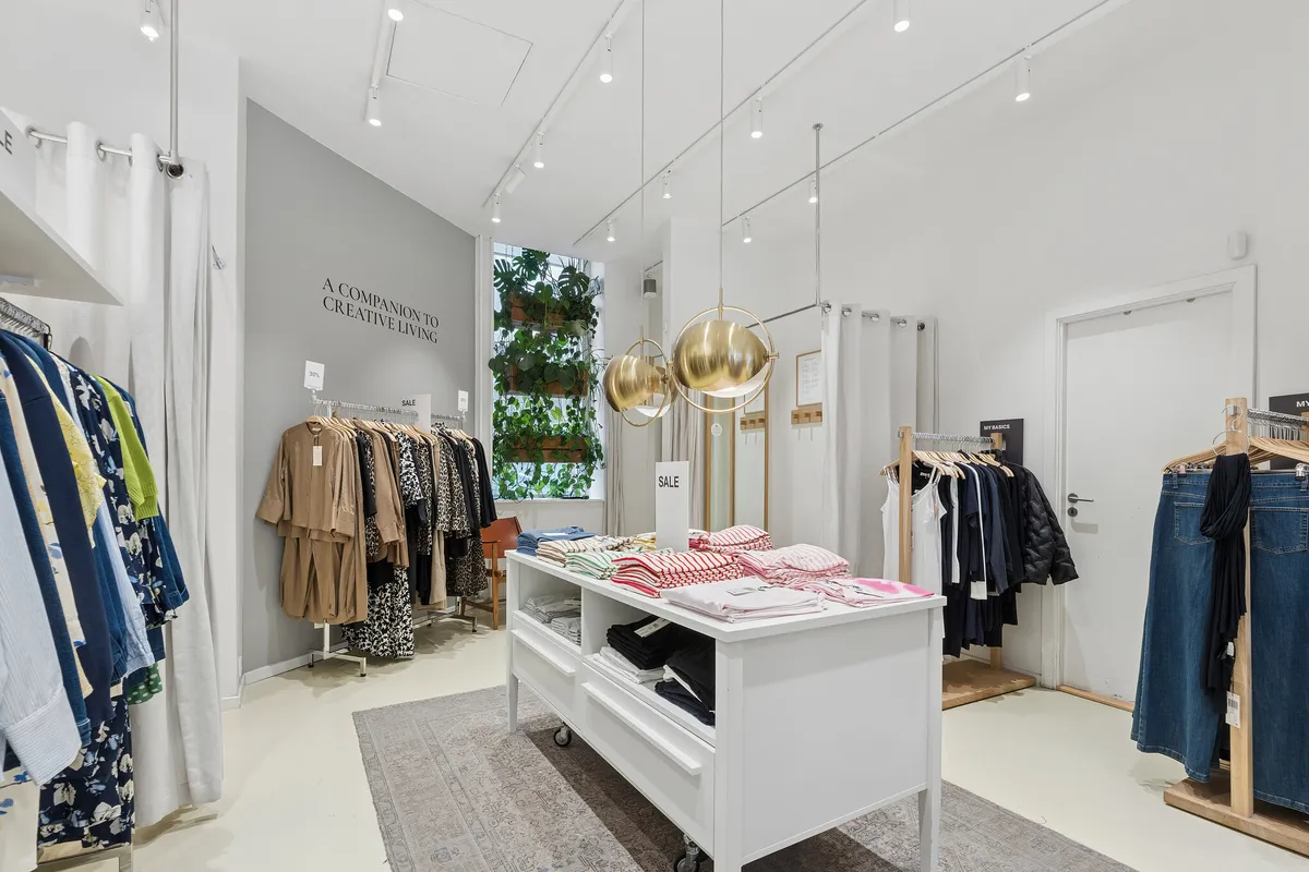 Et moderne butikslejemål med en stor, hvid midterbord, hvor tøj er udstillet og solgt. Rundt om er der hængehylder med tøj og trægulve. Der er hængende lysekroner og plantegrønne i baggrunden