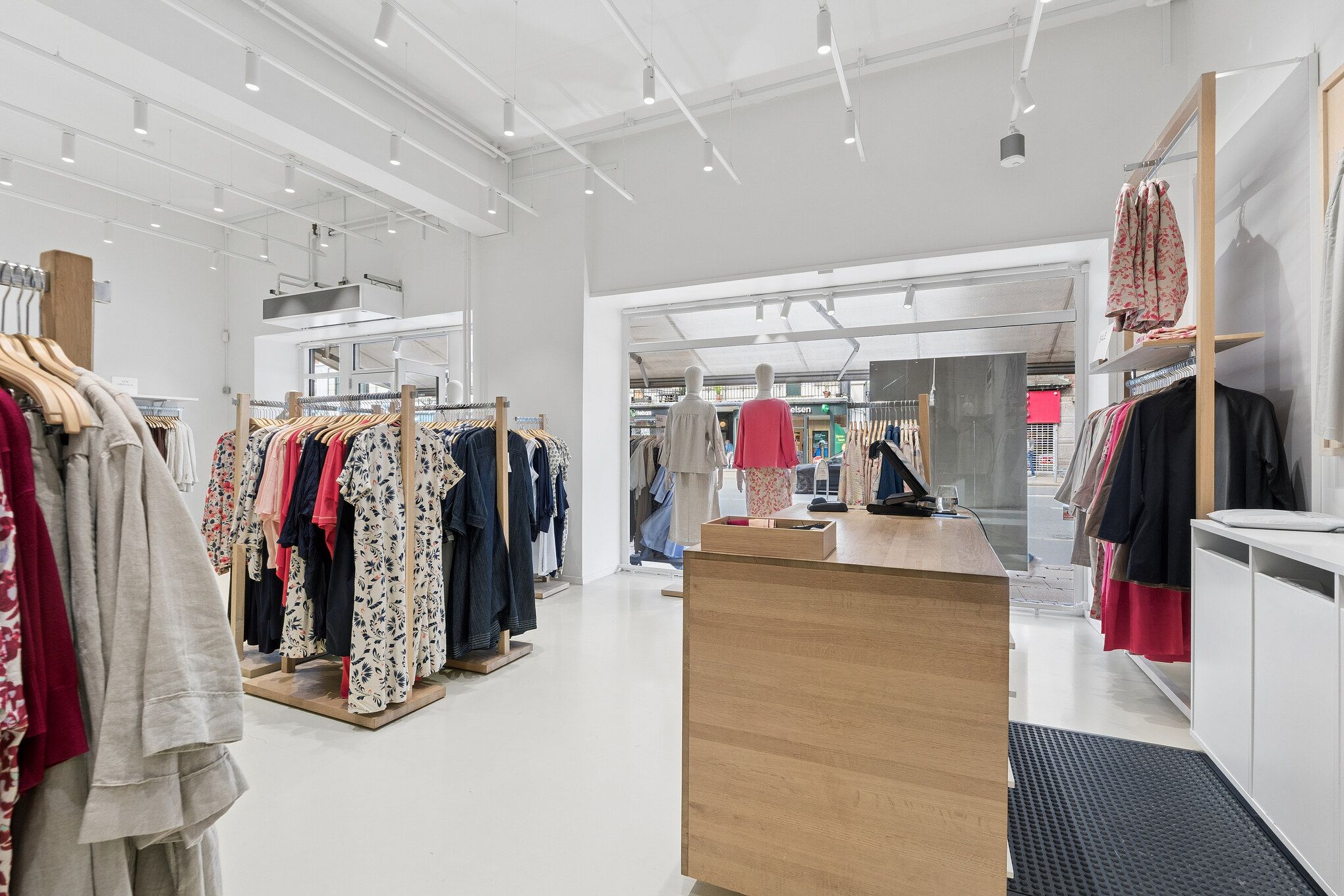 Modernt indrettet butikslokal med hvid og lys interiør og flotte klæder på tøjstativer og tøjstativ. Butik med stort glasfront og lysekroner i loftet.