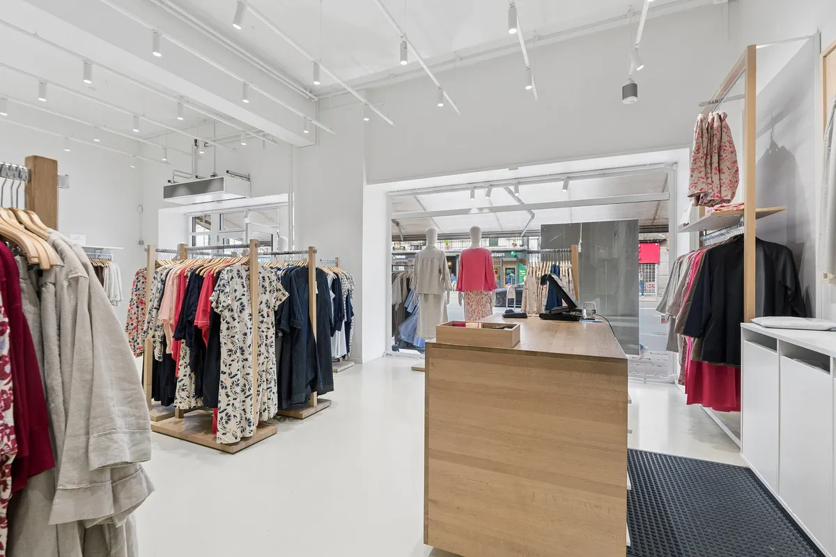 Modernt indrettet butikslokal med hvid og lys interiør og flotte klæder på tøjstativer og tøjstativ. Butik med stort glasfront og lysekroner i loftet.
