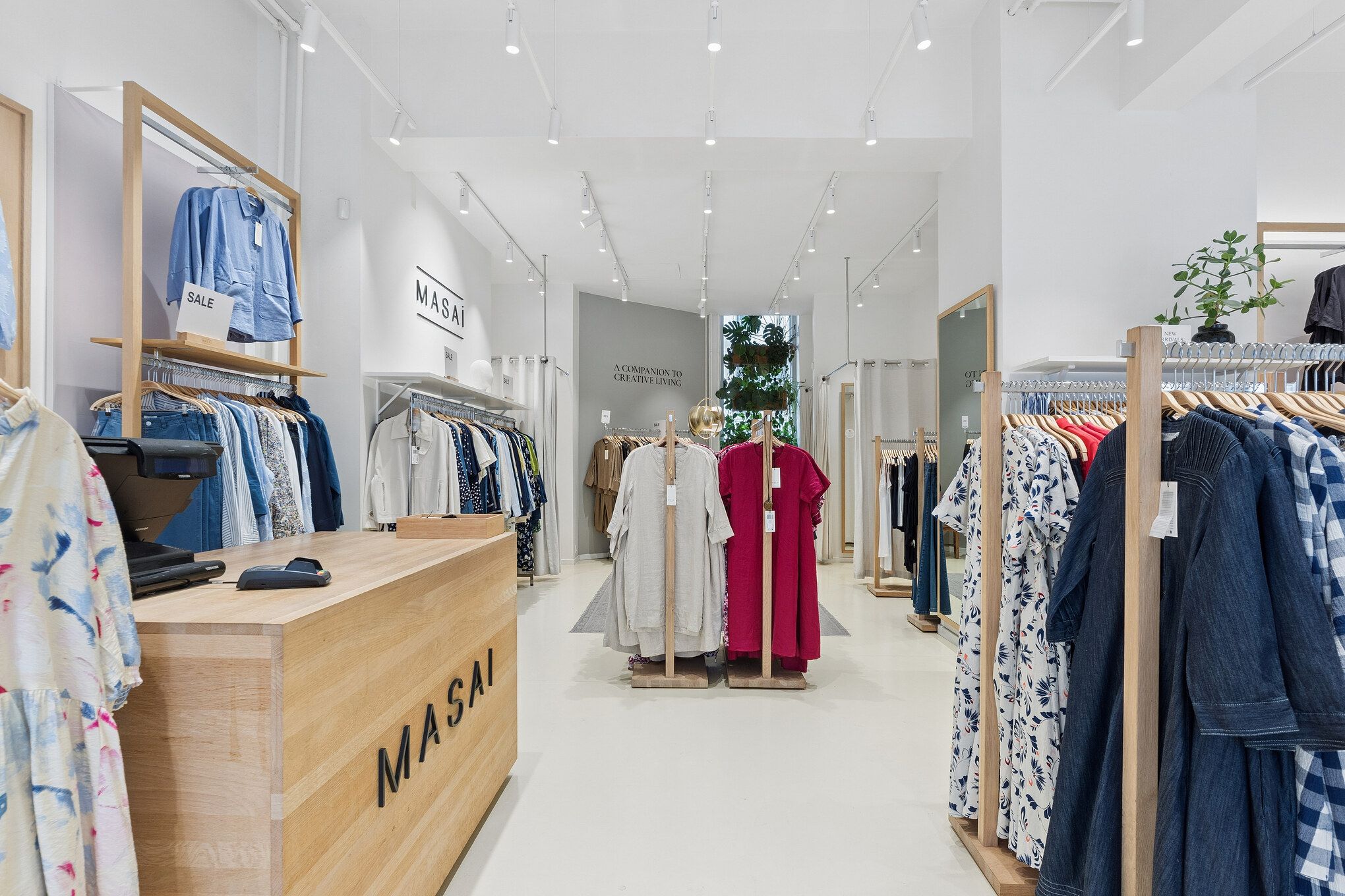 Butik med møbler i lyse træ og hvide vægge. Tøj på hængere og i højere hylder. En masse tøj i lyse farver og mønstre. Et bord med kasseapparat og mærkeskilt