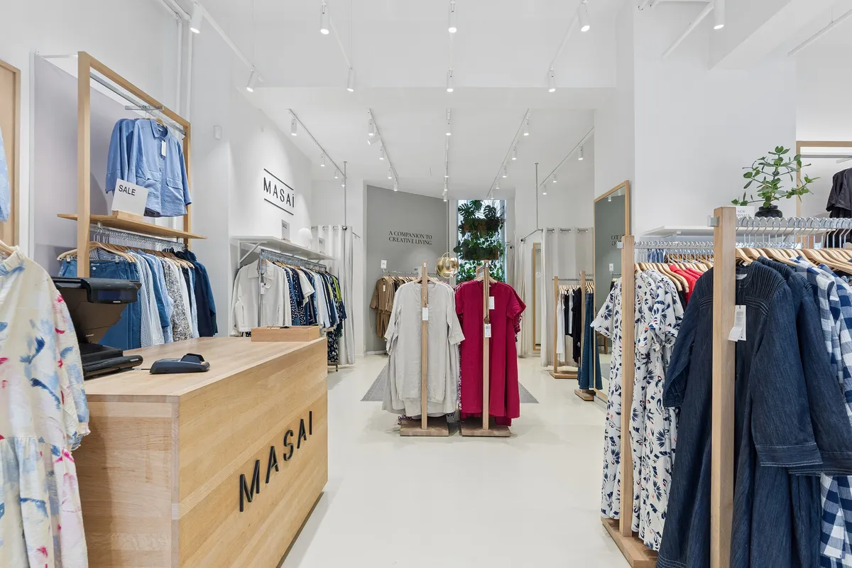 Butik med møbler i lyse træ og hvide vægge. Tøj på hængere og i højere hylder. En masse tøj i lyse farver og mønstre. Et bord med kasseapparat og mærkeskilt