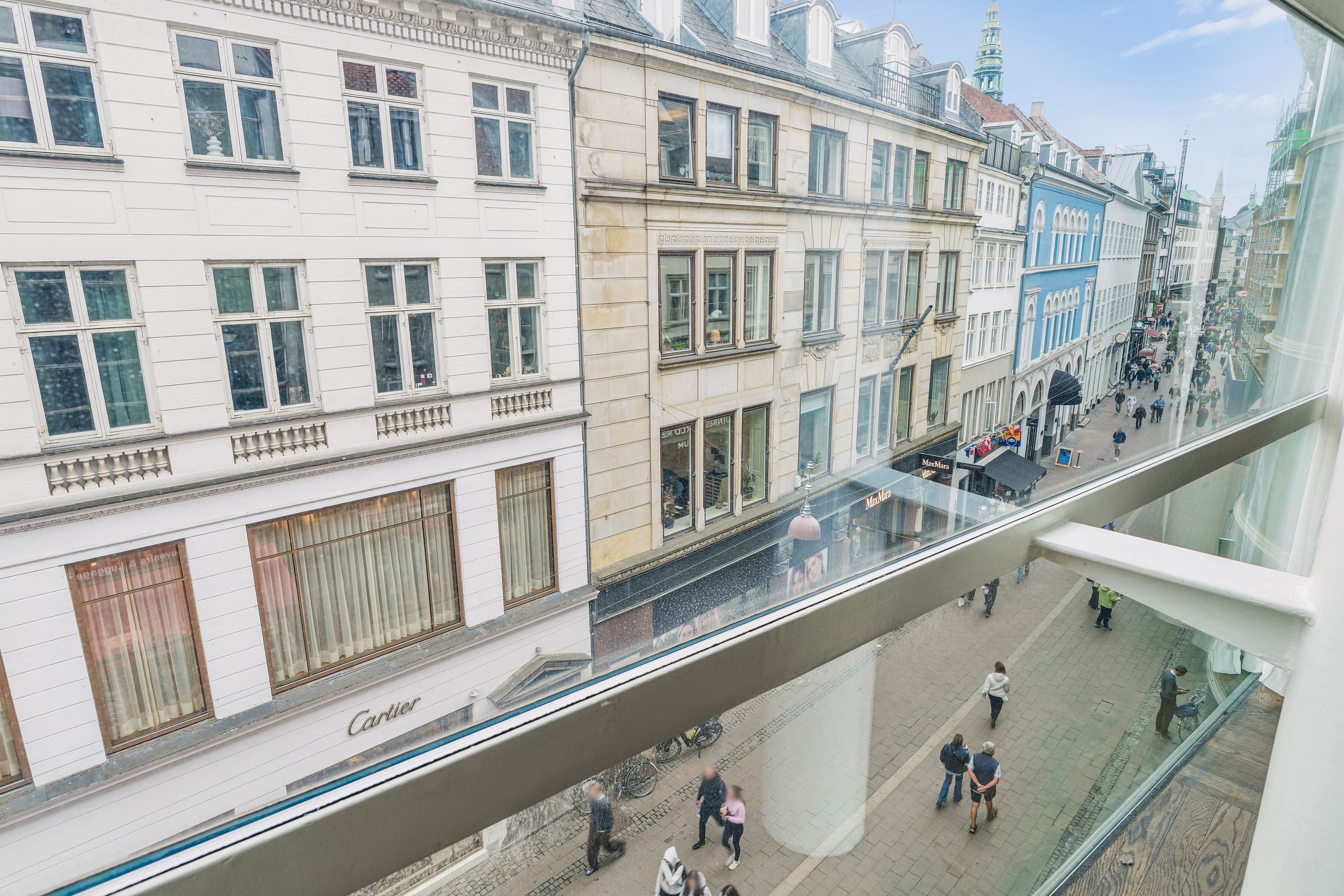 Fra et vindue ser man en gågade med mennesker, bygninger og en bil. På højre side er der en butik med skiltet Cartier. Bygningen er hvid og brun og er i centrum af København.