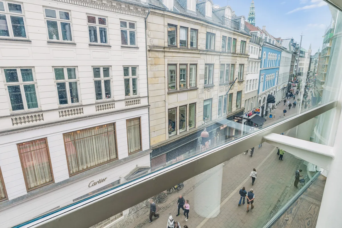Fra et vindue ser man en gågade med mennesker, bygninger og en bil. På højre side er der en butik med skiltet Cartier. Bygningen er hvid og brun og er i centrum af København.