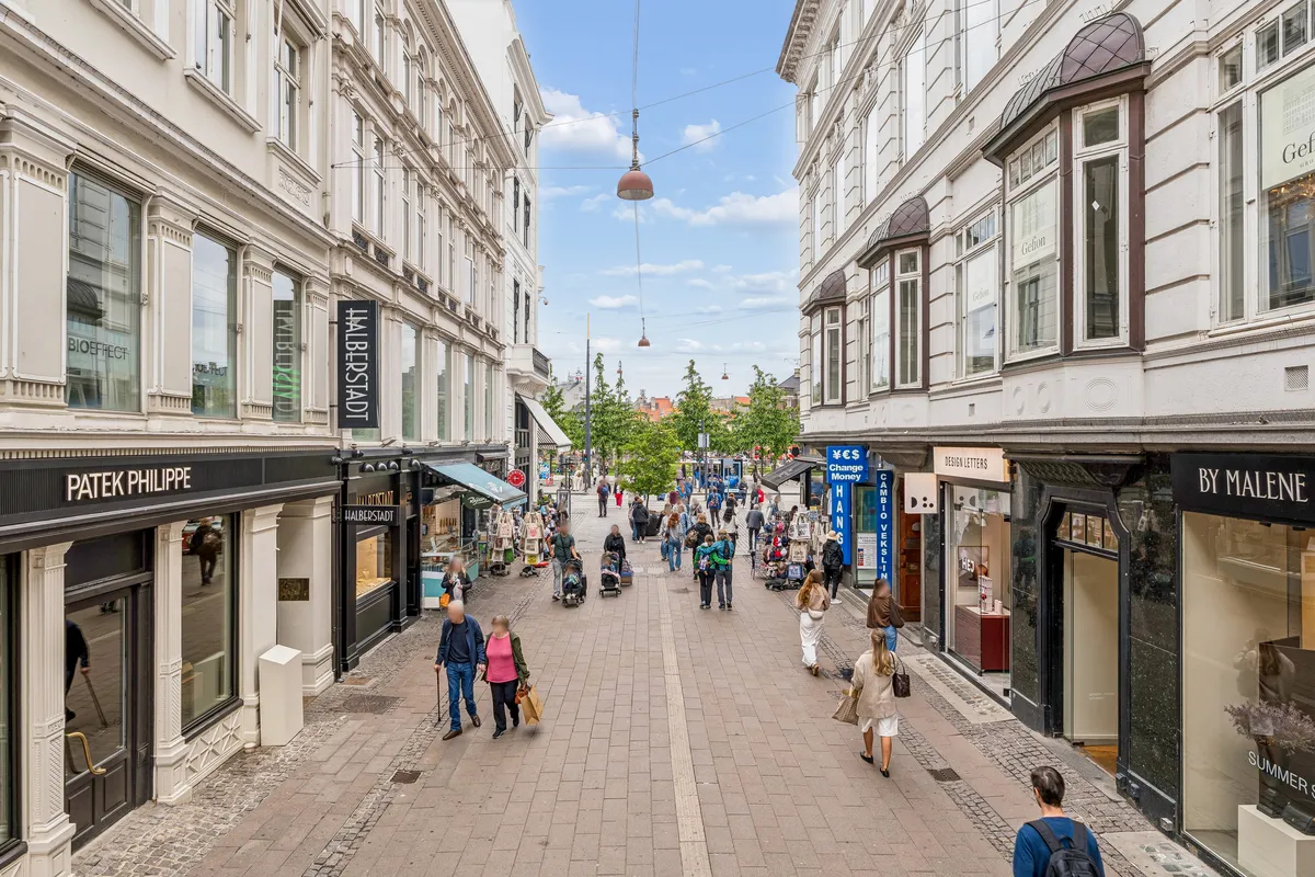 På Østergade i København ser vi mennesker, der går, og butikker med navne som Patek Philippe og By Malene. En guldhandel og en smykkeforretning er også synlige.