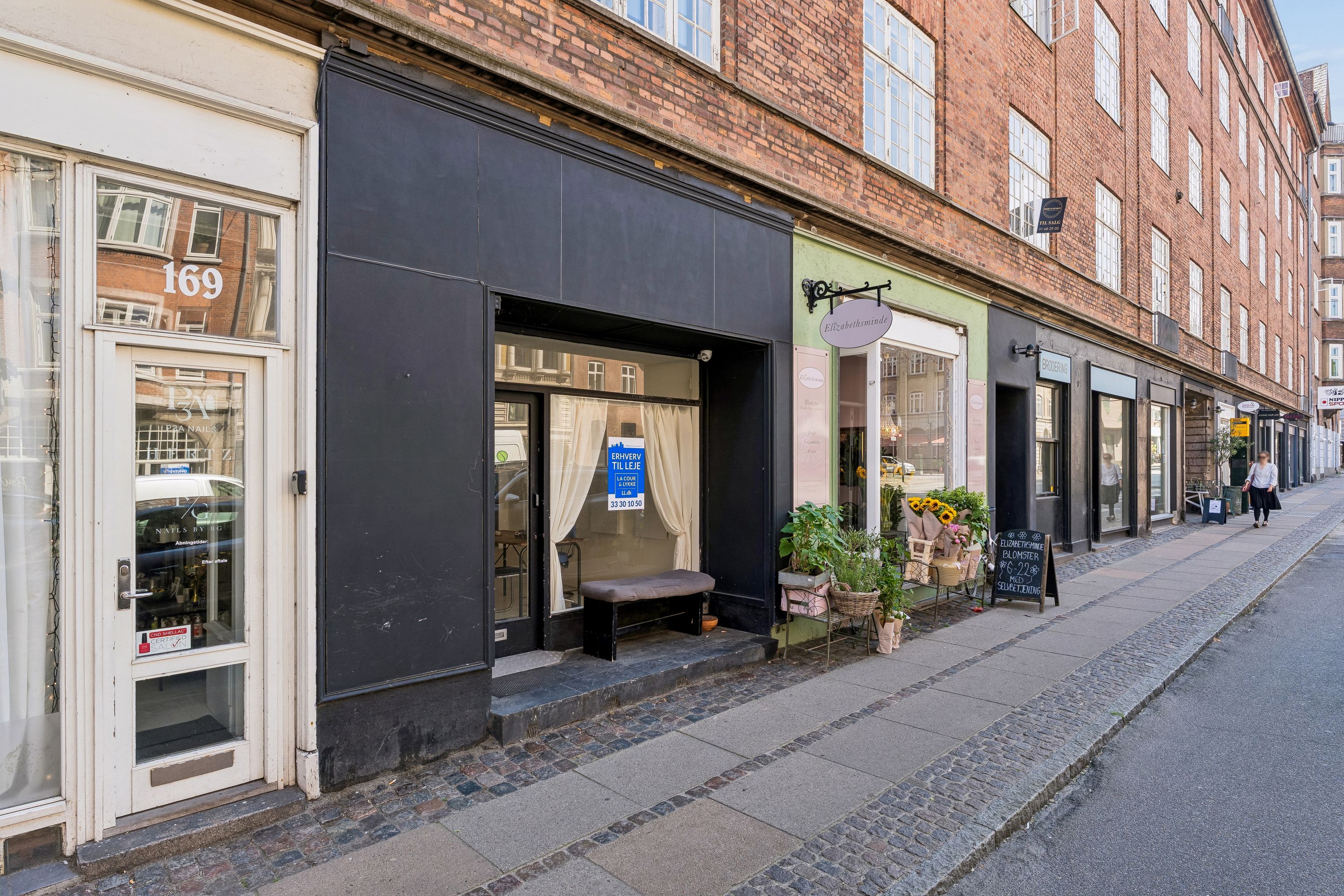 Butik med udsalgsvinduer på Vesterbrogade 169 i Frederiksberg, med plantager og blomster på fortovet og 2 personer, der går på fortovet