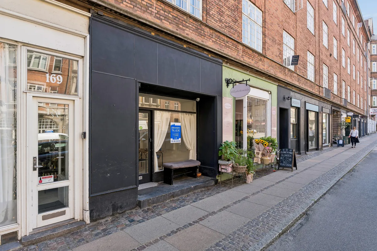 Butik med udsalgsvinduer på Vesterbrogade 169 i Frederiksberg, med plantager og blomster på fortovet og 2 personer, der går på fortovet