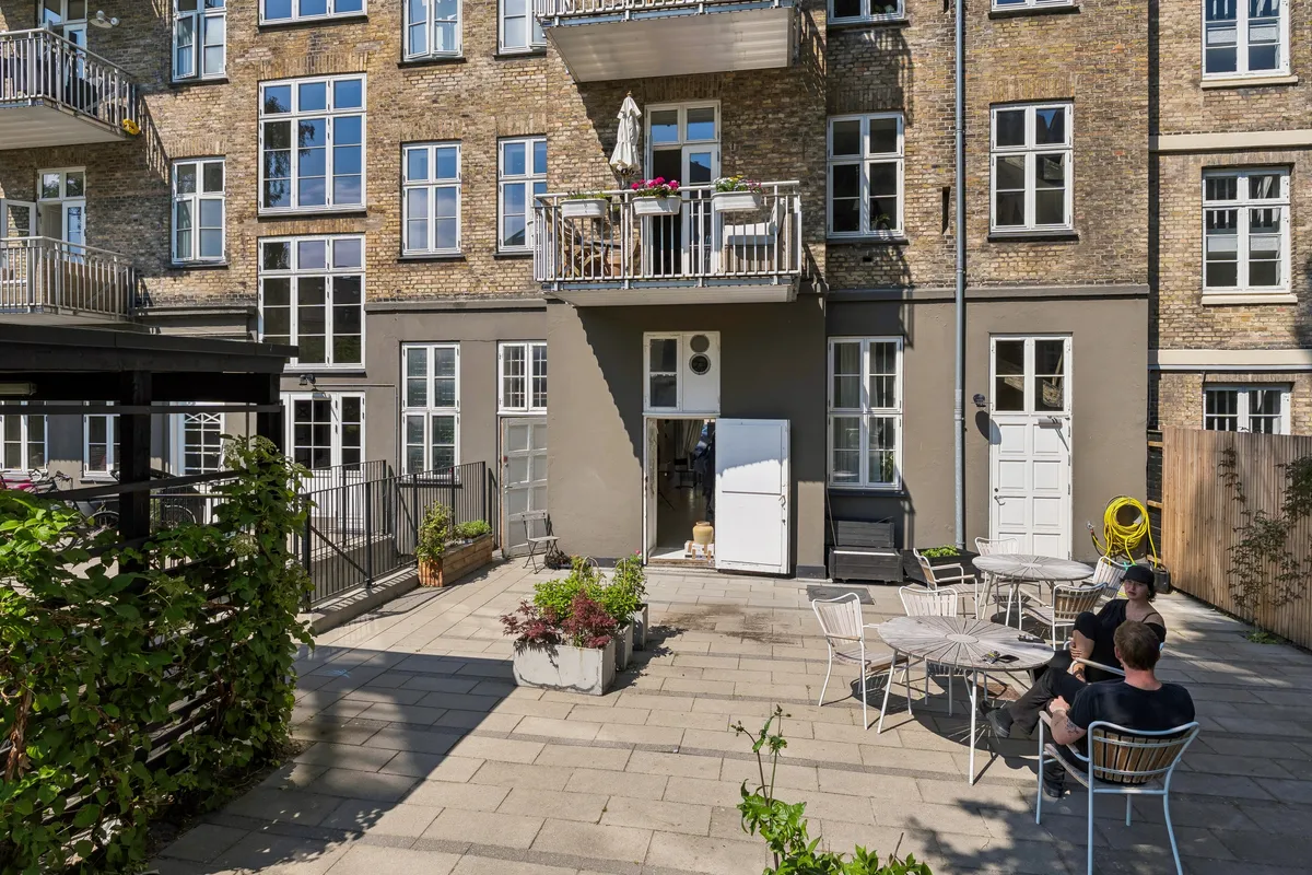 To personer sidder og hviler sig på en terrasse foran en bygning med flere etager og balkoner med blomster.