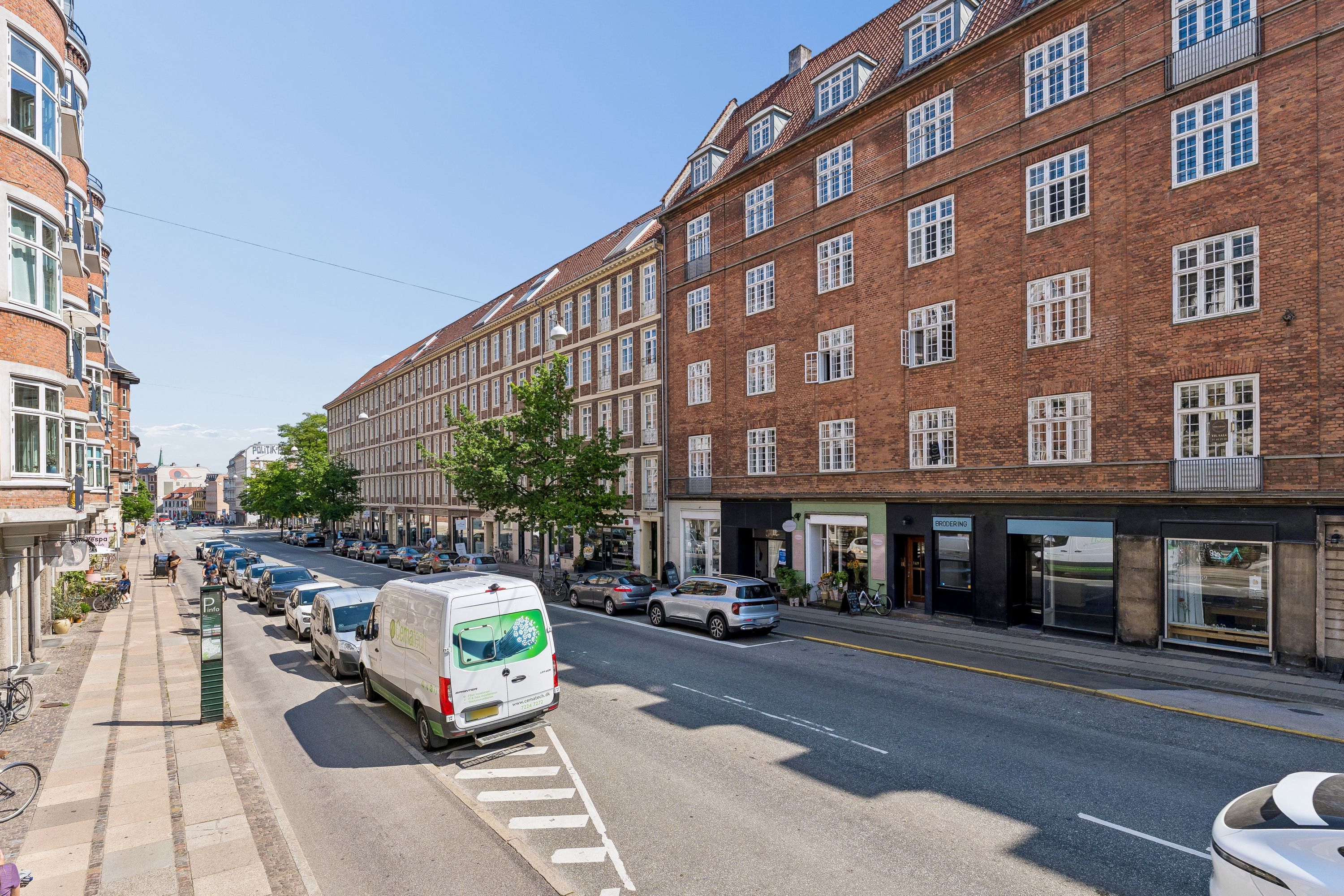 Mennesker går langs gaden med parkerede biler, der er flere butikker og bygninger med vinduer langs gaden