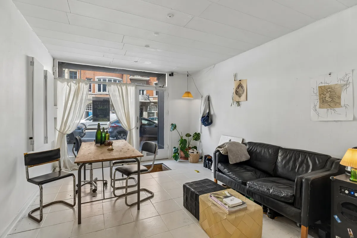 Lejlighed med åbent køkken og stue med lyse hvide vægge og møbler, hvor der er et gult stolpelys og en sort sofa med et bord og et skur.