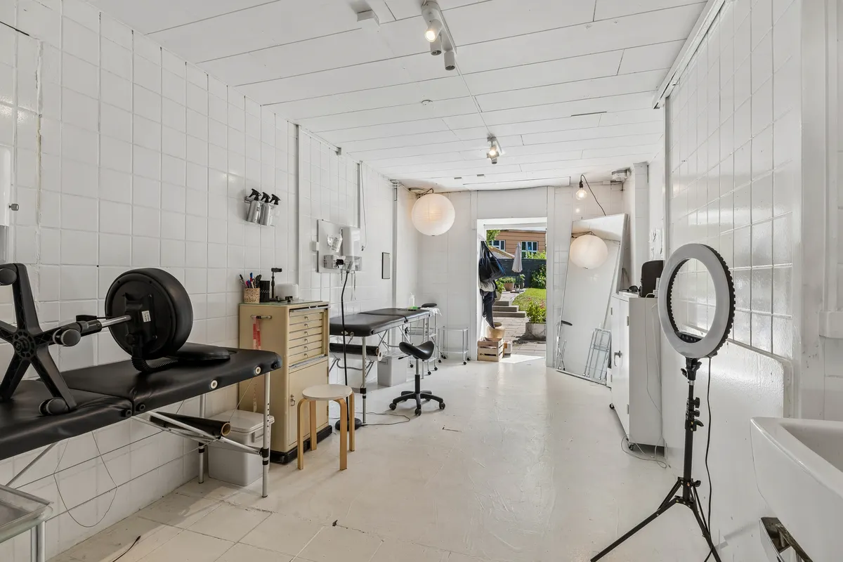Inde i en hvidmalet lægepraksis er der udstyr som en massagestol, en behandlingsstol og en rund spejl. Der er også en stålampe og et hvidt badekar.