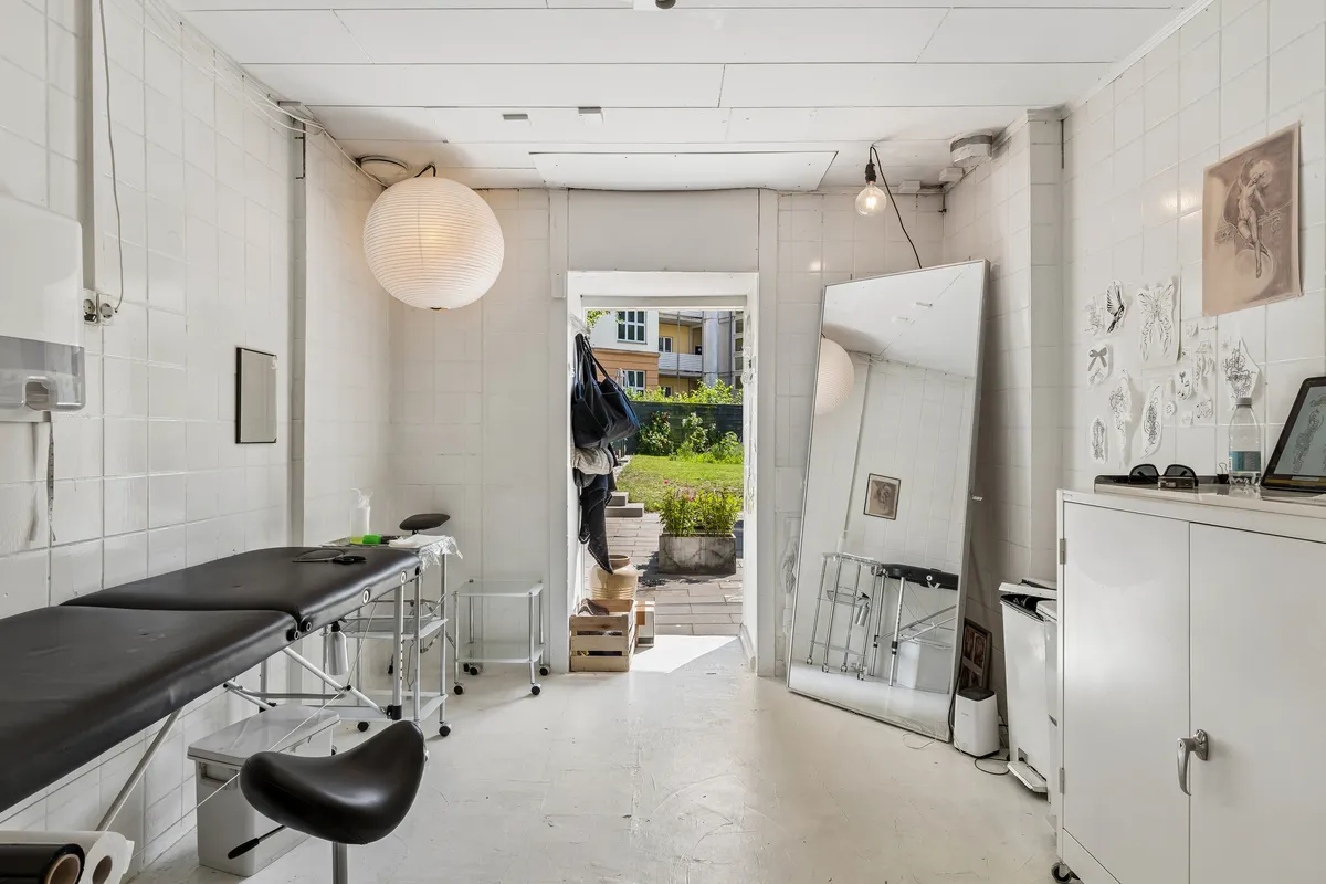 En hvid og minimalistisk massagesalon med træborde, høje hvide vægge og en stor spejl. Der er også en åben dør med udsigt til en have med planter og grønne planter.