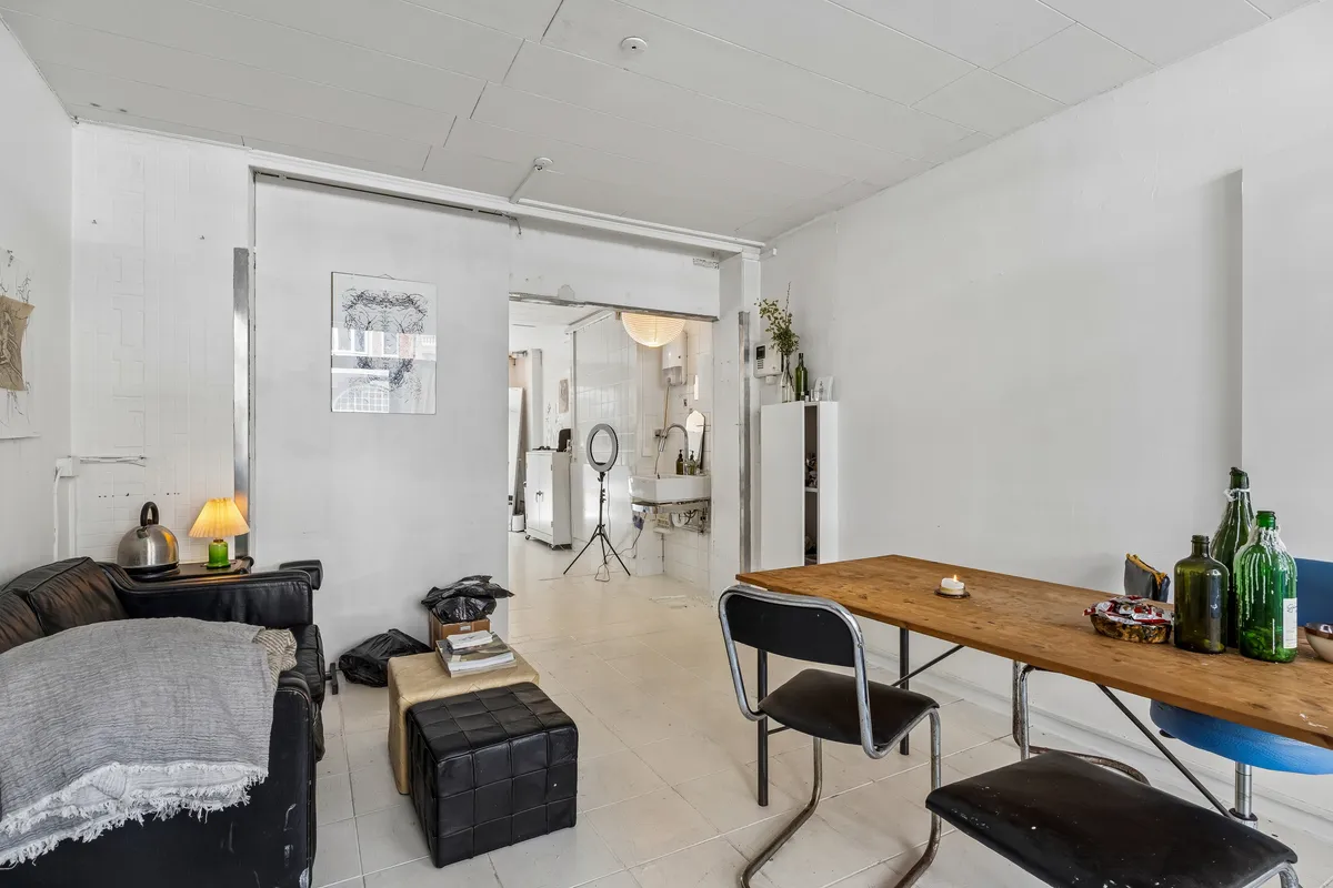 Butikslokale med sort sofa, gult bord, to stolpestole og en lampe samt en hvid køleskab og et bad med spejl i baggrunden