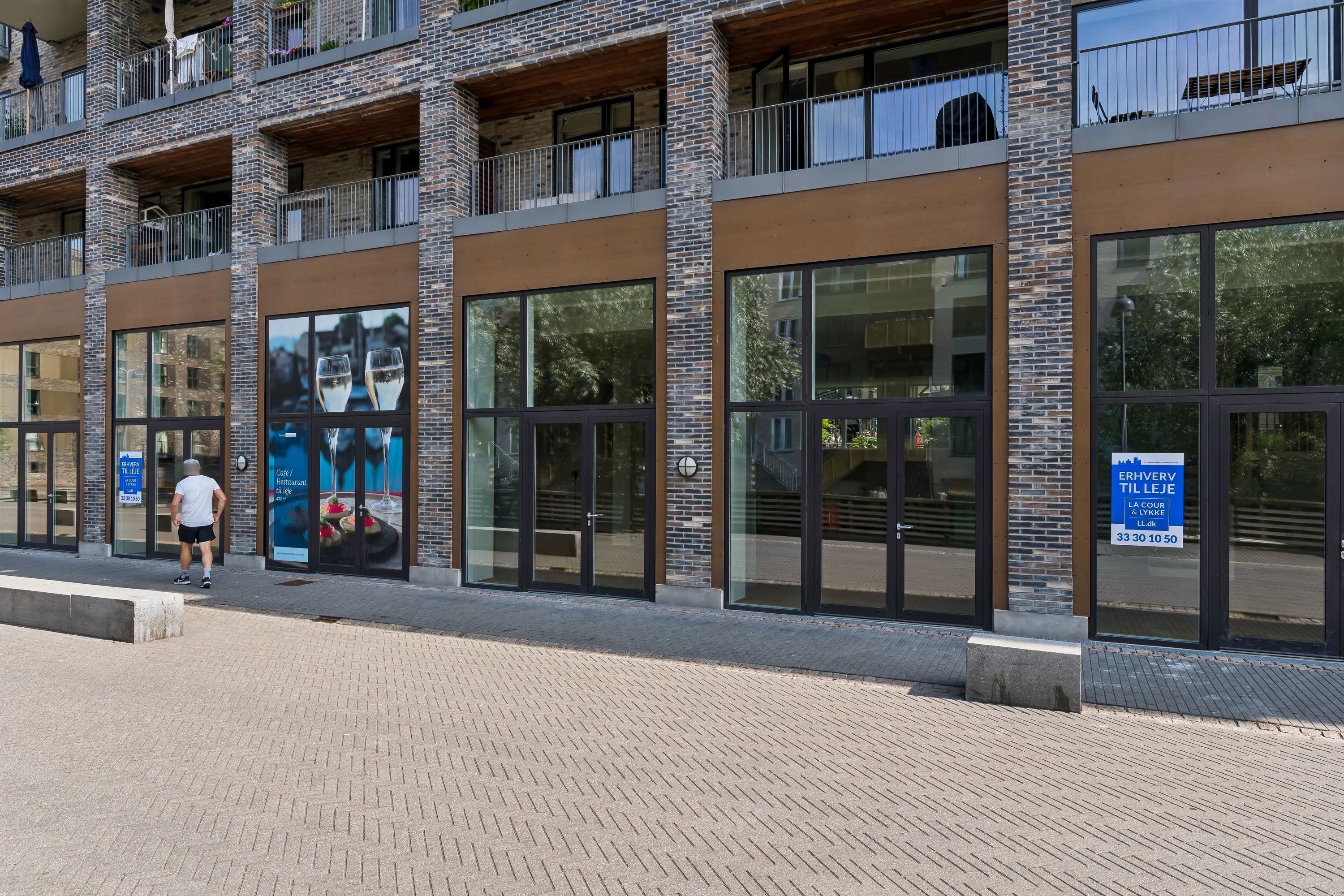 En mand går forbi en bygning med store vinduer, et søjledesign og en væg med møbler, sofaer og en stol. Der er et skilt med teksten "Erhover til leje" og en adresse.