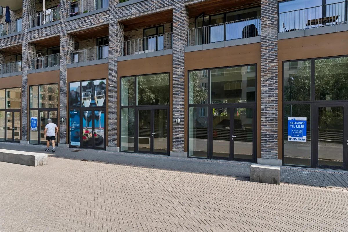En mand går forbi en bygning med store vinduer, et søjledesign og en væg med møbler, sofaer og en stol. Der er et skilt med teksten "Erhover til leje" og en adresse.