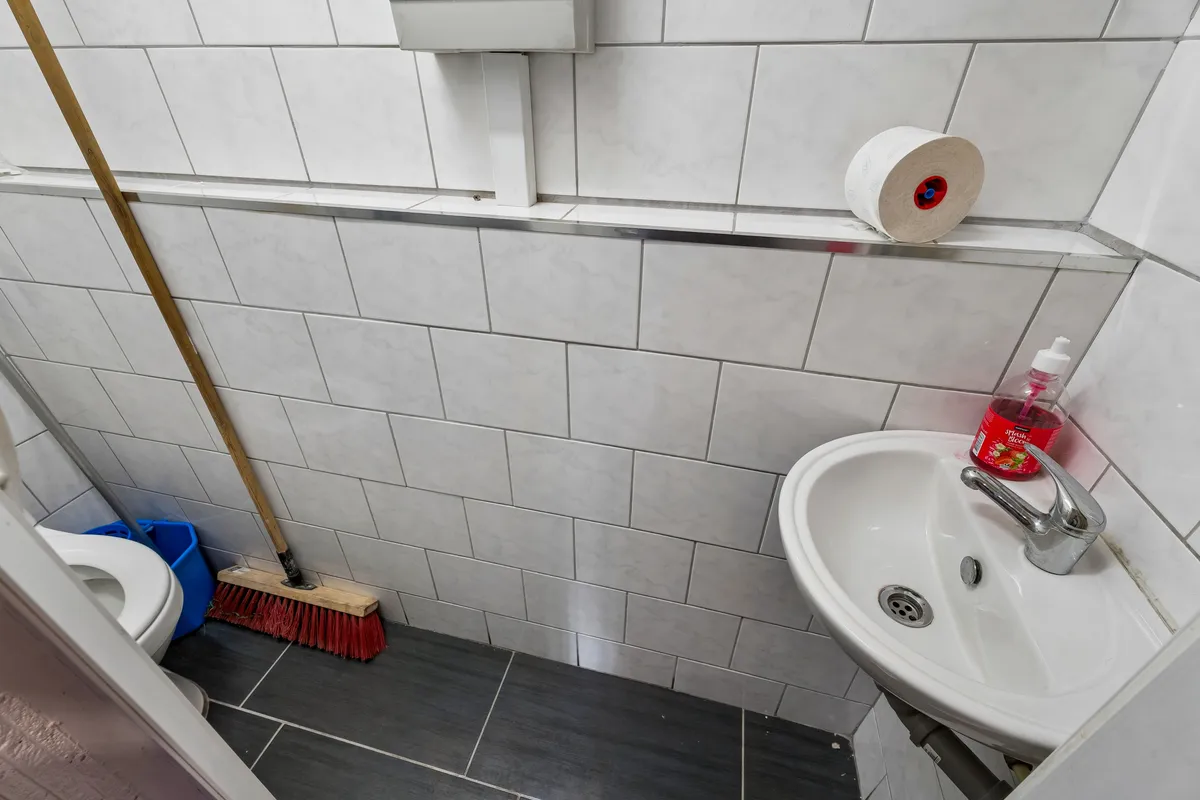 Badeværelse med hvide fliser, en toiletbørste, et toilet, et paprulle og en håndvask med en vandhane og håndsæbe.