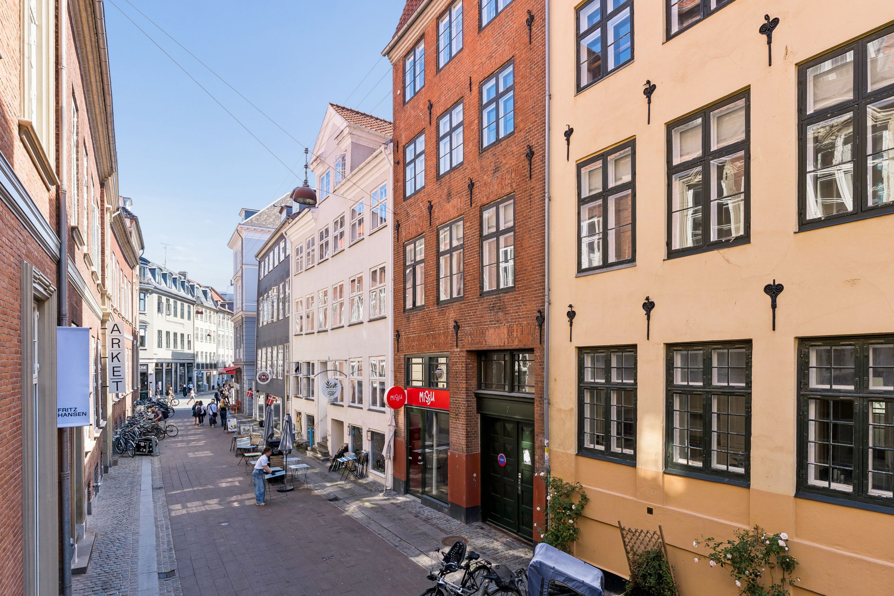 En smuk dansk bygade med røde og gule murstensbygninger, glasvinduer og folk som går og står langs gaden, med en bycykelparkering og en café med navnet MØSLØV.
