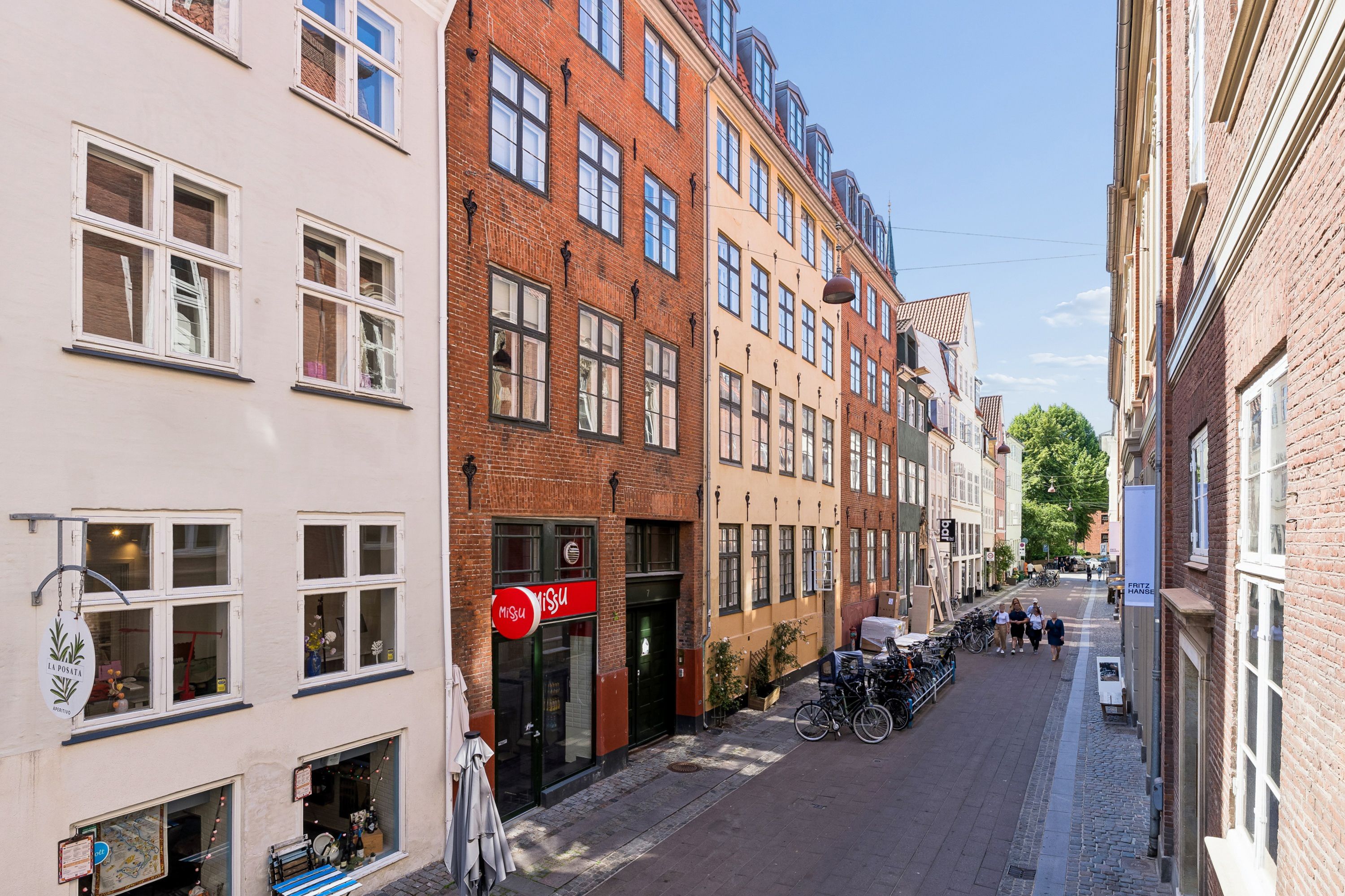 Et smukt ældre kvarter med bygninger i røde og gule mursten og hvide vinduerne og en bred gågade med mennesker, cykler og en facade med "Miso"-skilt
