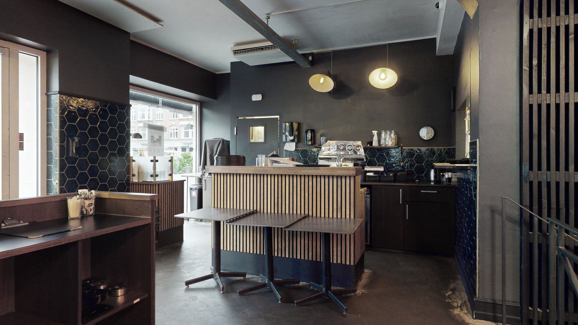Moderne og elegant køkken med mørke vægge og lyse loftslampe. Værelse med to høje stolpe og køkkenudstyr i baggrunden. Træborde med lyse loftslamper i midten og et hegn mod højre side.