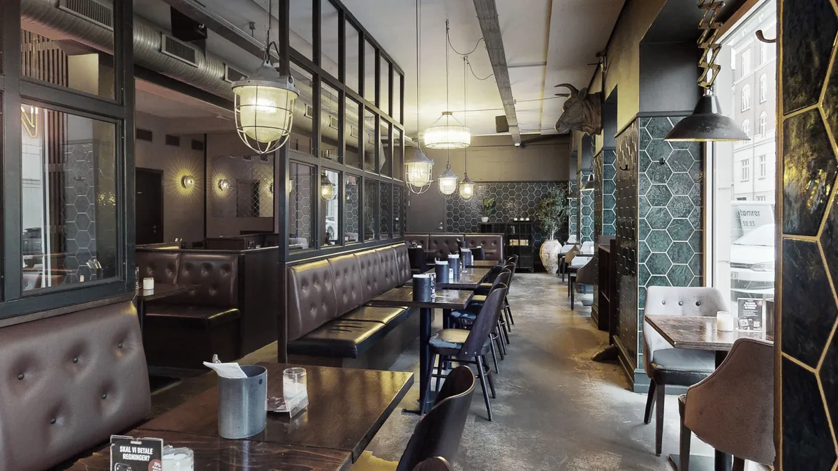 Køkkenbord med lyse lamper, lædersofaer, træborde og stolpe med kvæghovde og lyse gulvbelægning i en mørk restaurant med heksagonmønstre og lyse vinduer