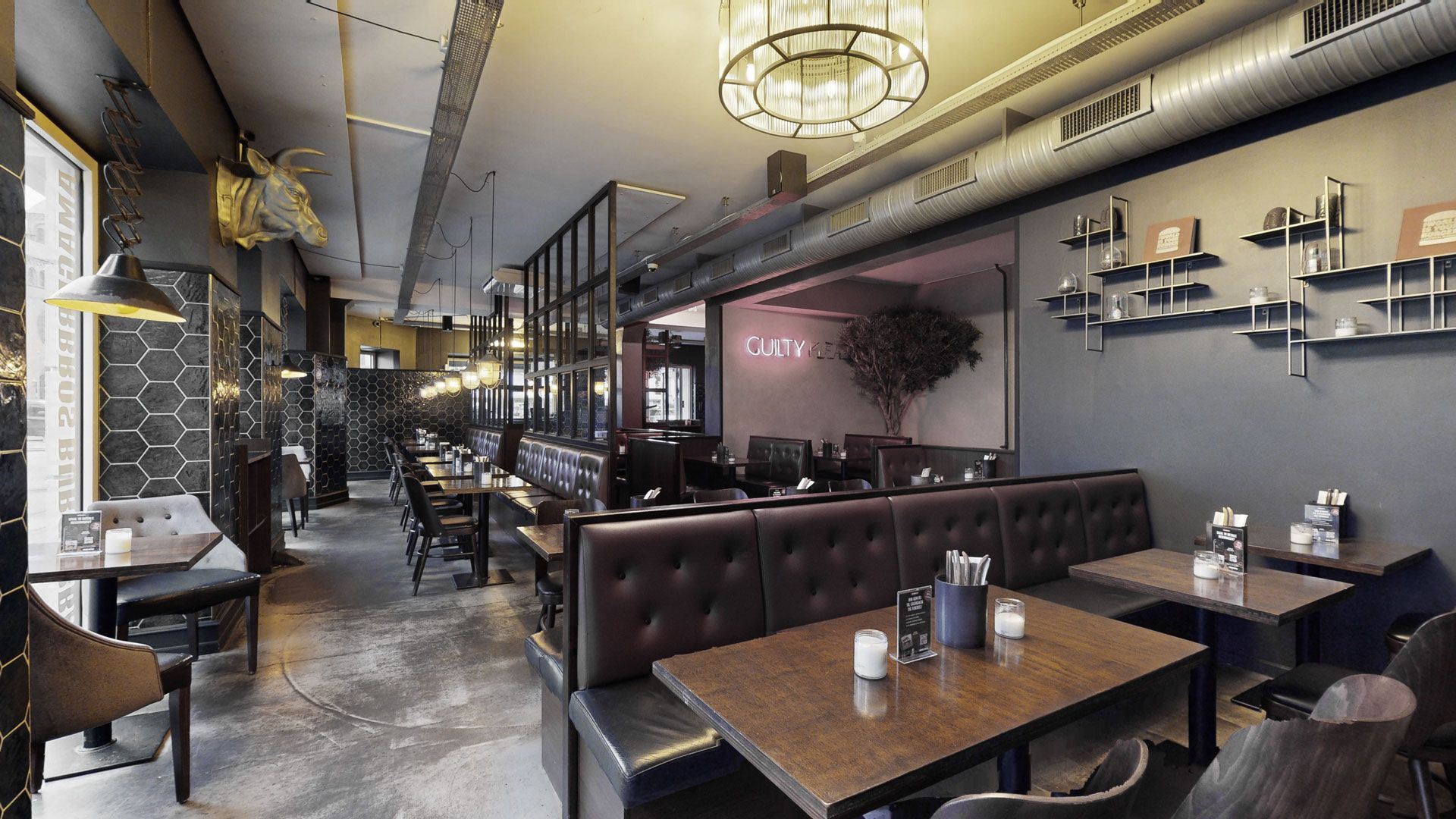 Rummeligt restaurantlejemål med højt loft, store vinduer, mørke læderstole og lyse loftslamper samt en gul neonskilt med teksten "Guilty"